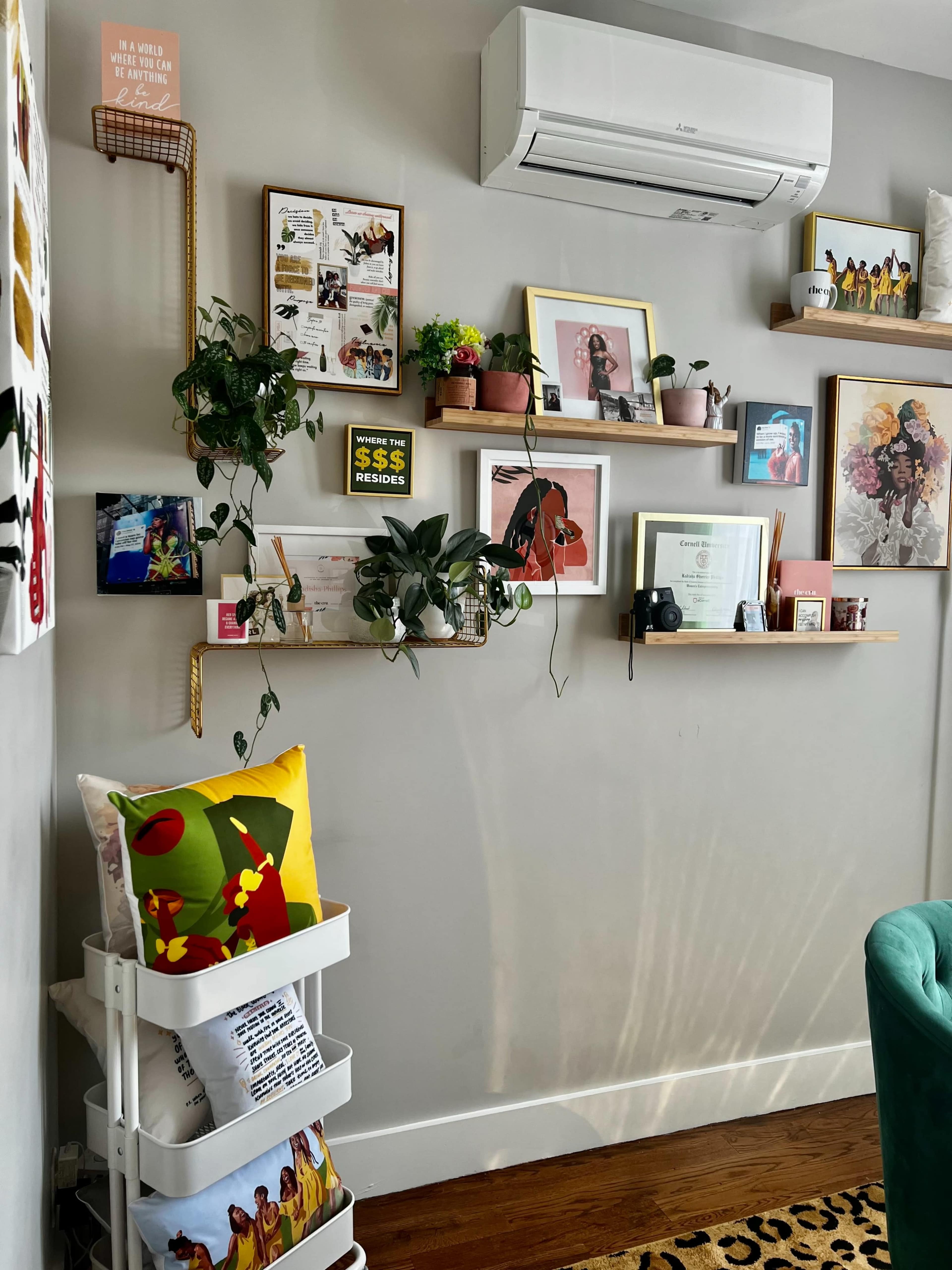 The image shows a cozy wall in a room adorned with various framed pictures, plants on shelves, and a small cart holding decorative pillows and items.
