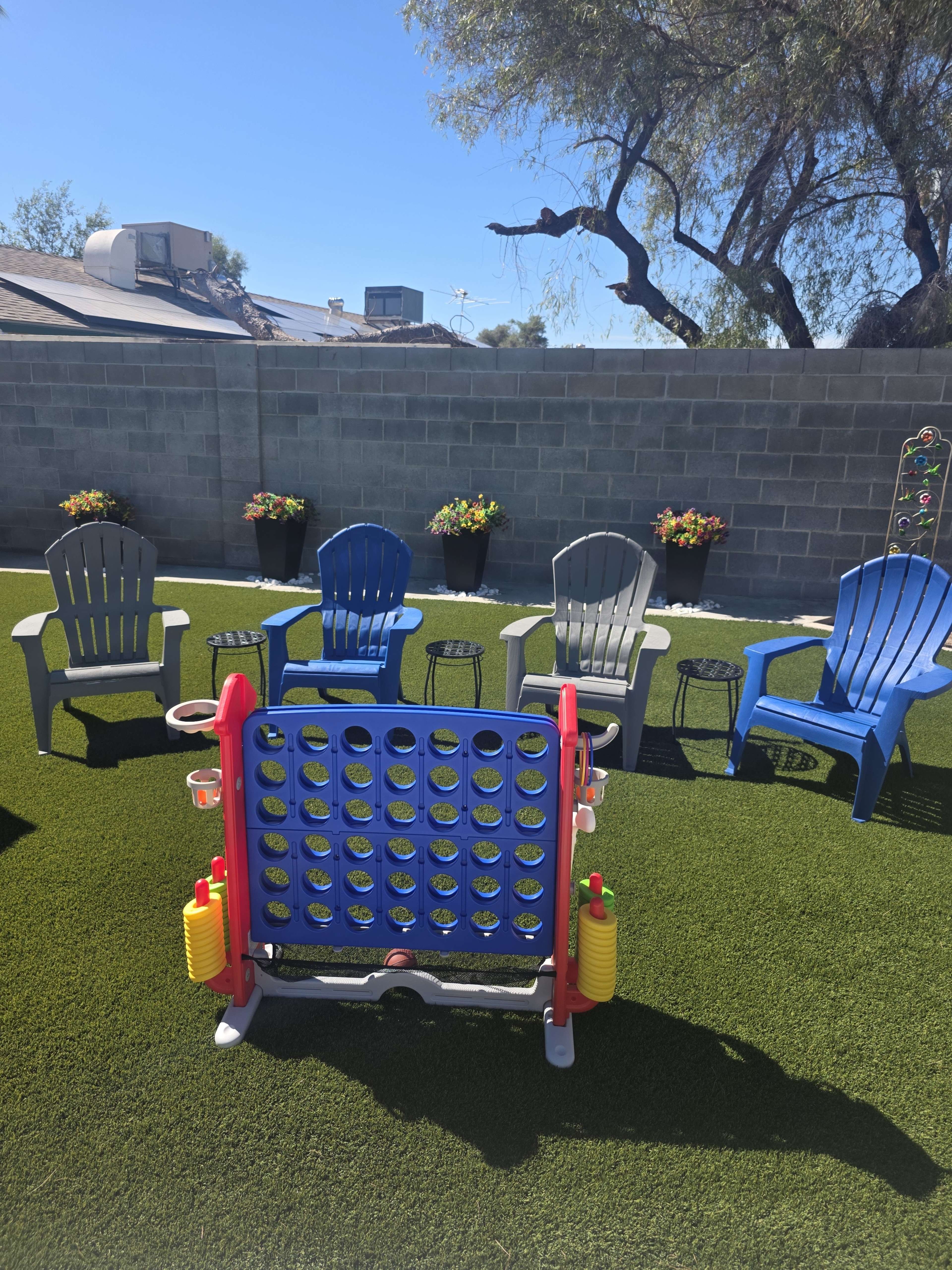 The image shows a backyard area with several colorful chairs arranged around a large Connect Four game set on artificial grass.