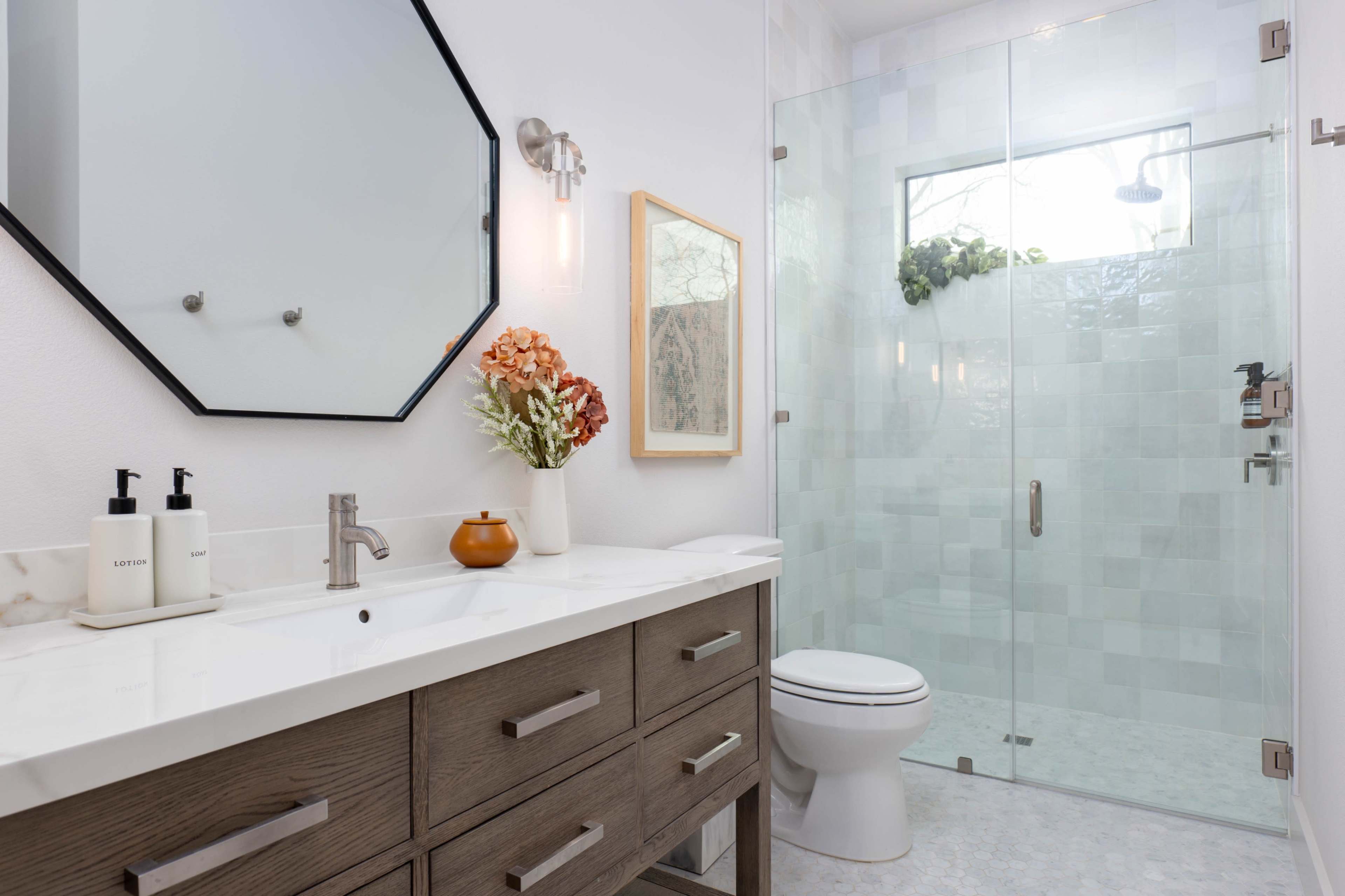The image shows a modern bathroom featuring a glass shower, a wooden vanity with a white countertop, and a decorative wall mirror.