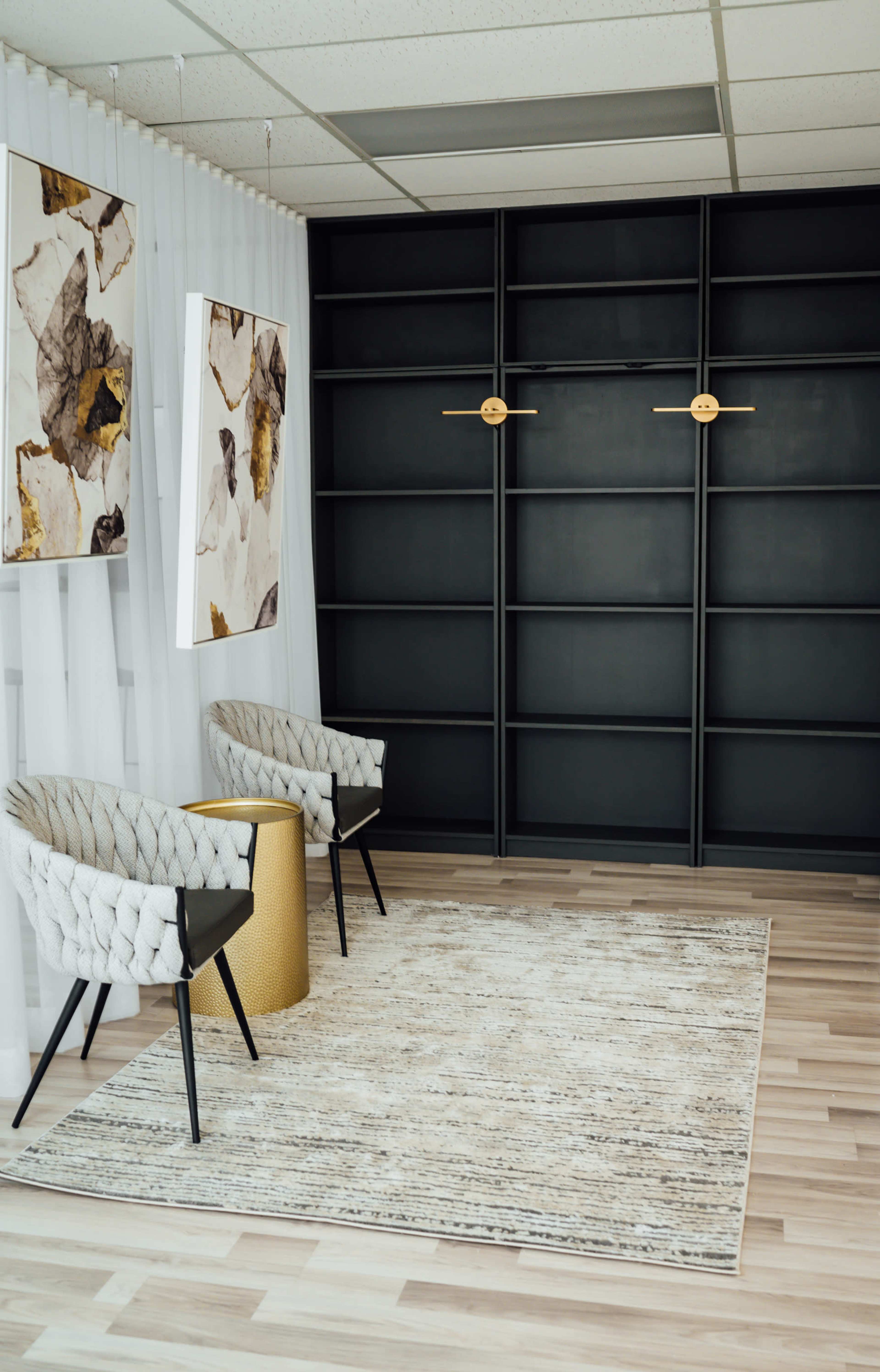 A minimalist interior features two patterned chairs, a round gold table, and abstract artwork hanging on the walls, alongside a large black shelving unit.