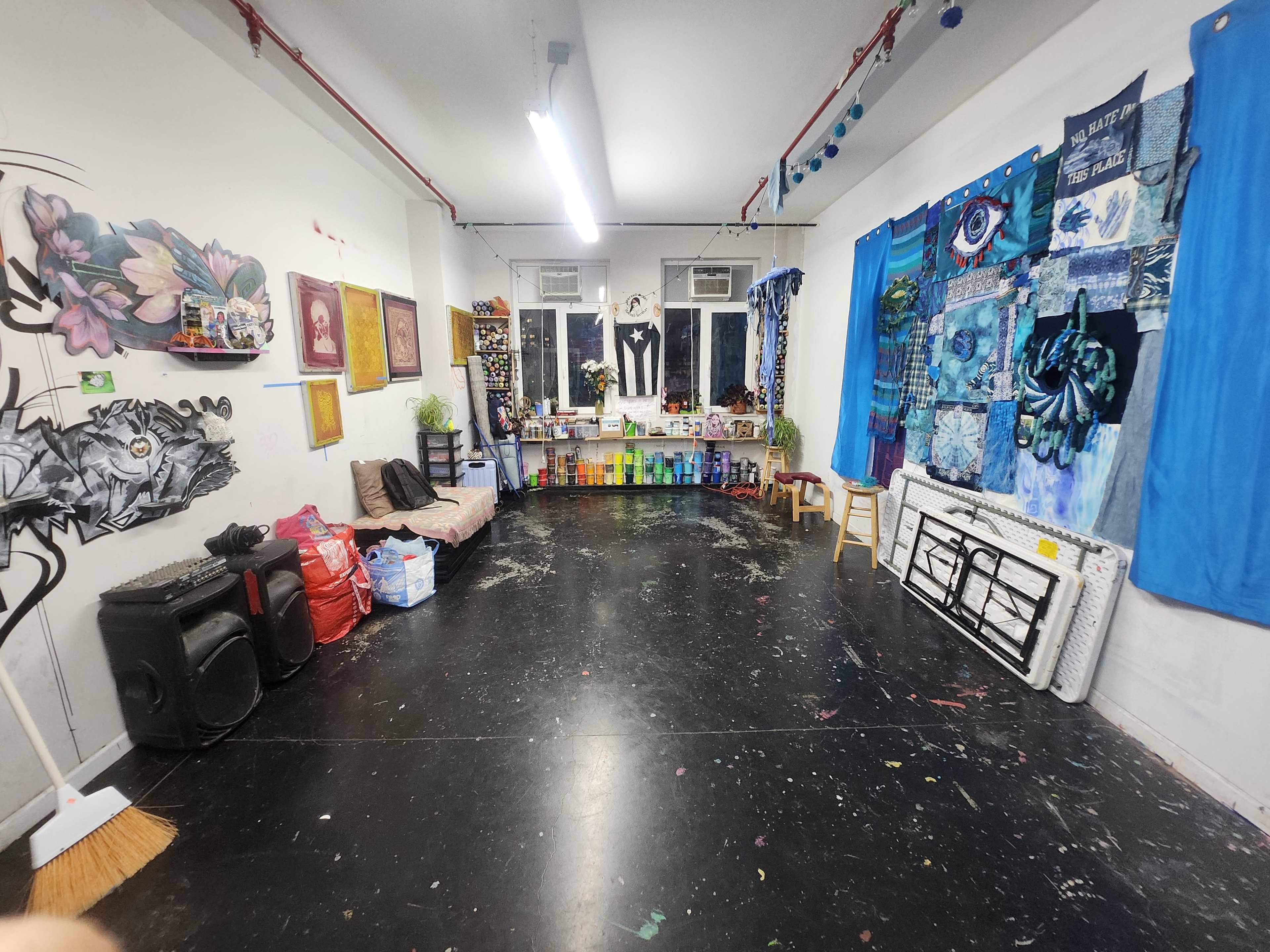 The image shows a spacious art studio with various artworks on the walls, a seating area with a couch, and a cluttered counter displaying art supplies and decorations.