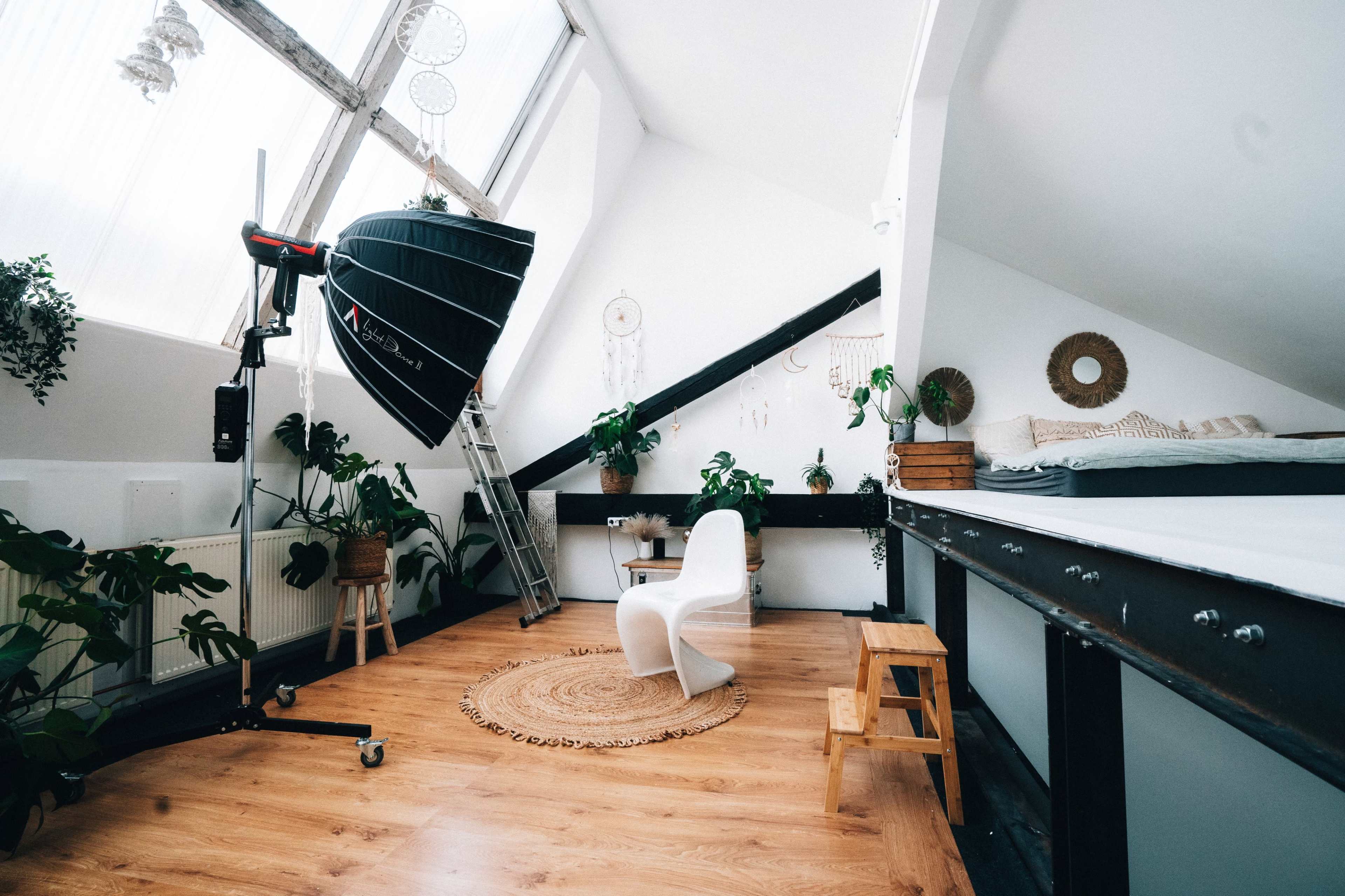 A well-lit attic studio with wooden flooring, a white chair, a large photography light, and various plants along the walls.