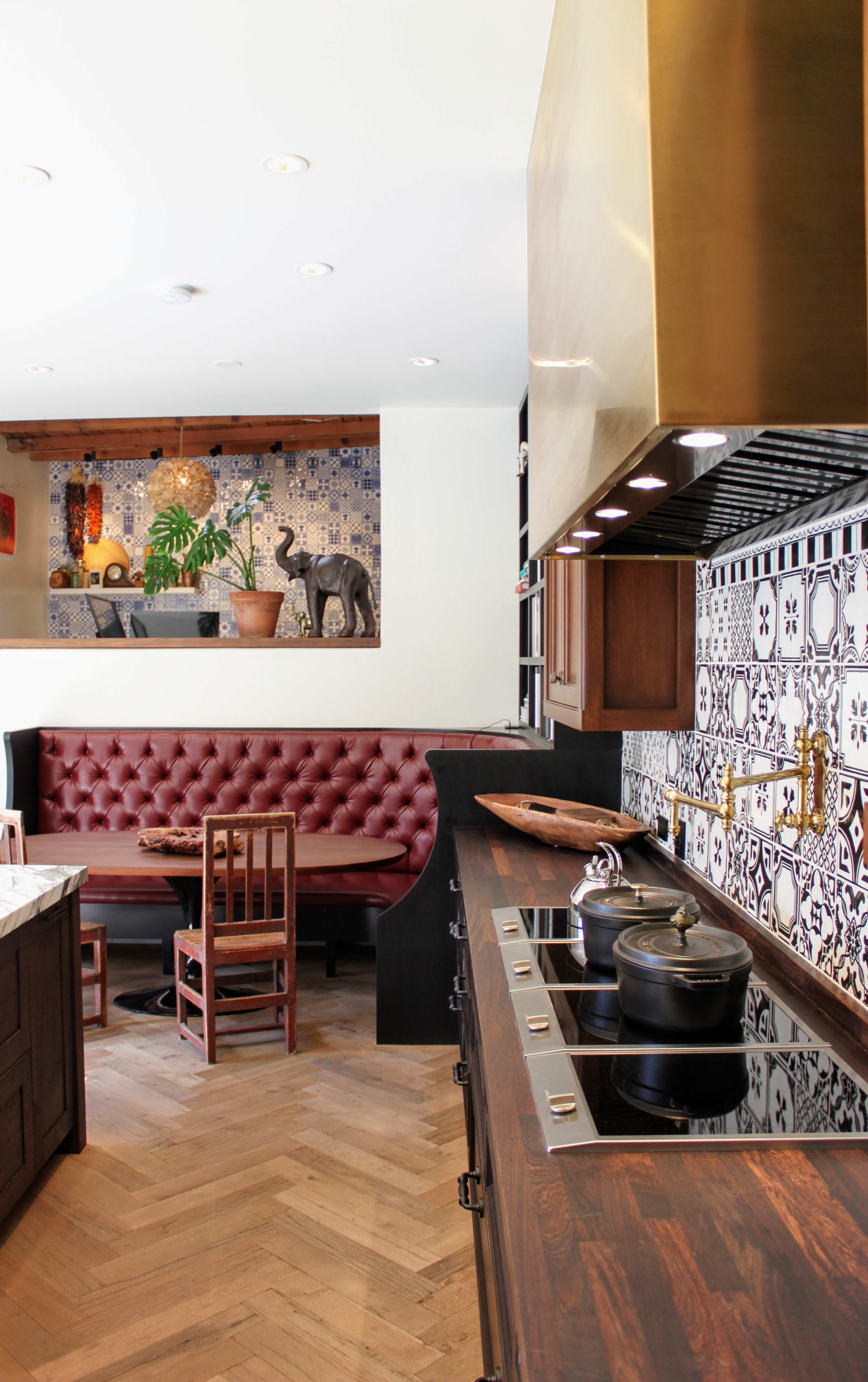 A modern kitchen features a gold range hood, an intricate tiled backsplash, and a cozy dining area with a burgundy banquette and wooden chairs.