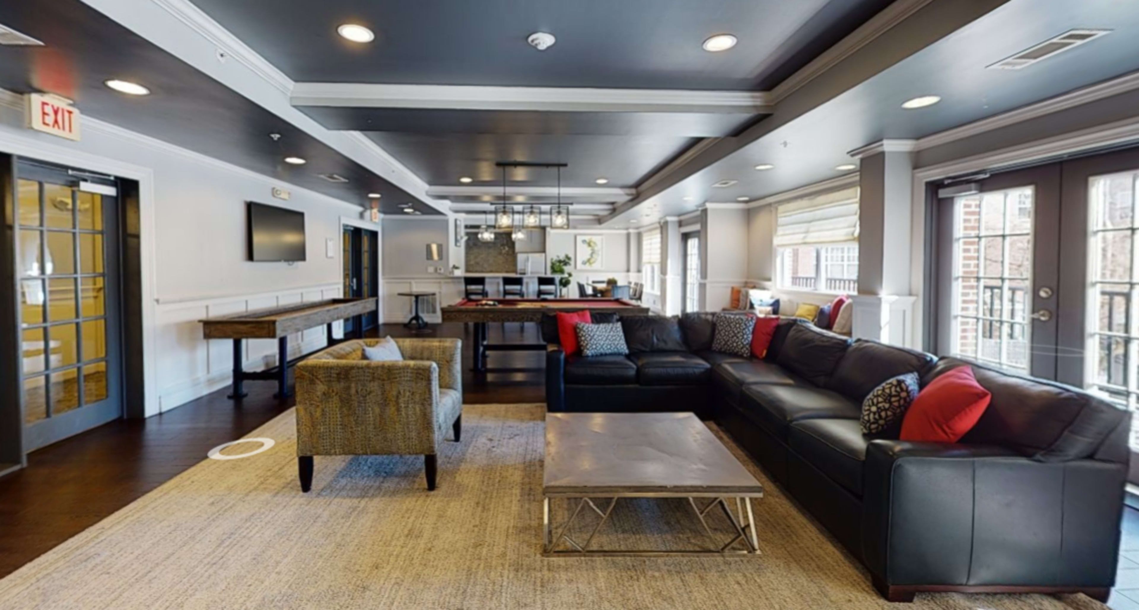 A modern lounge area with a black leather sectional sofa, a wooden table, and large windows, featuring a view of a kitchen and dining space.