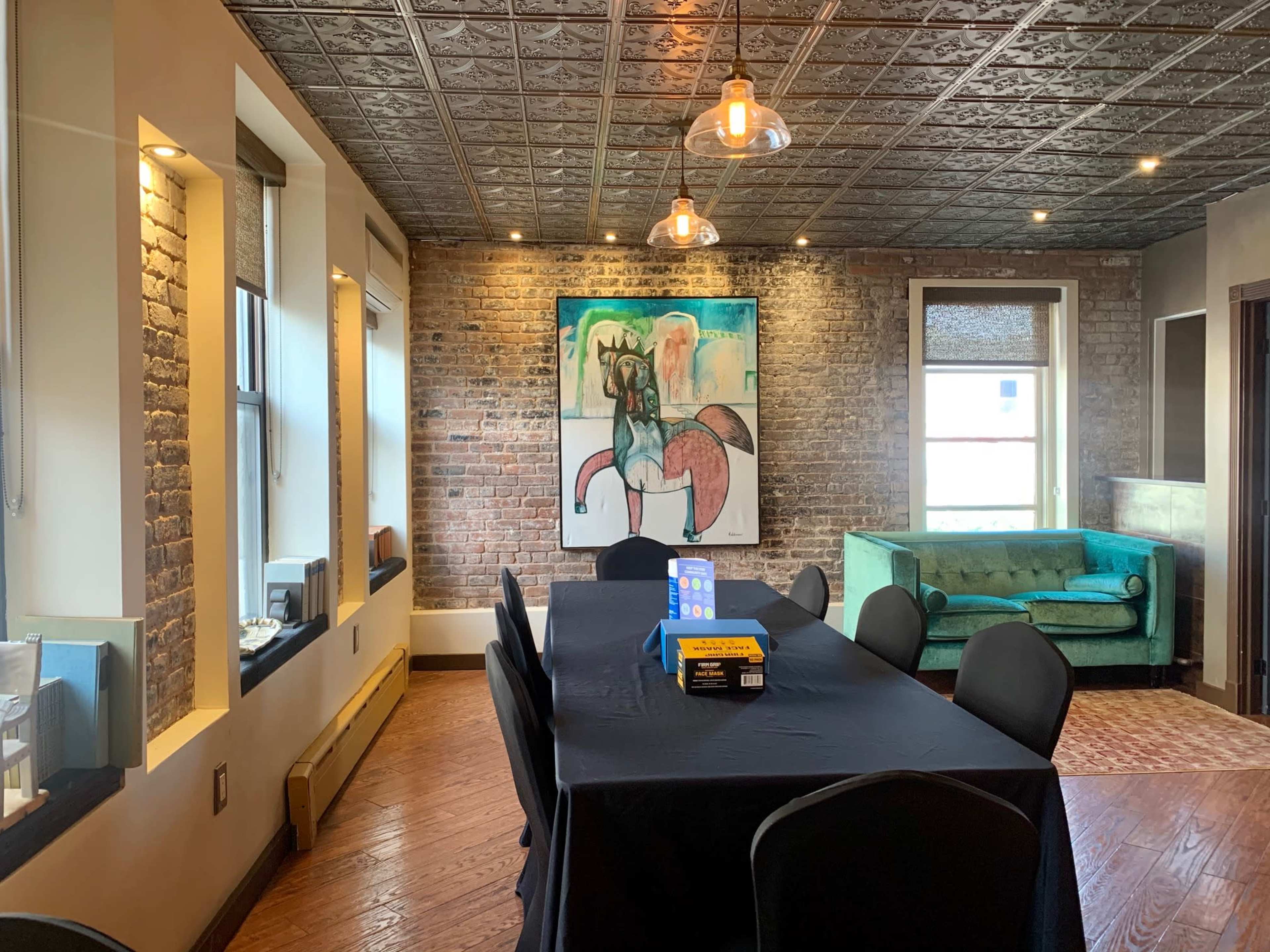 A modern meeting room with a large black table, black chairs, a green sofa, exposed brick walls, and a colorful artwork on the wall.