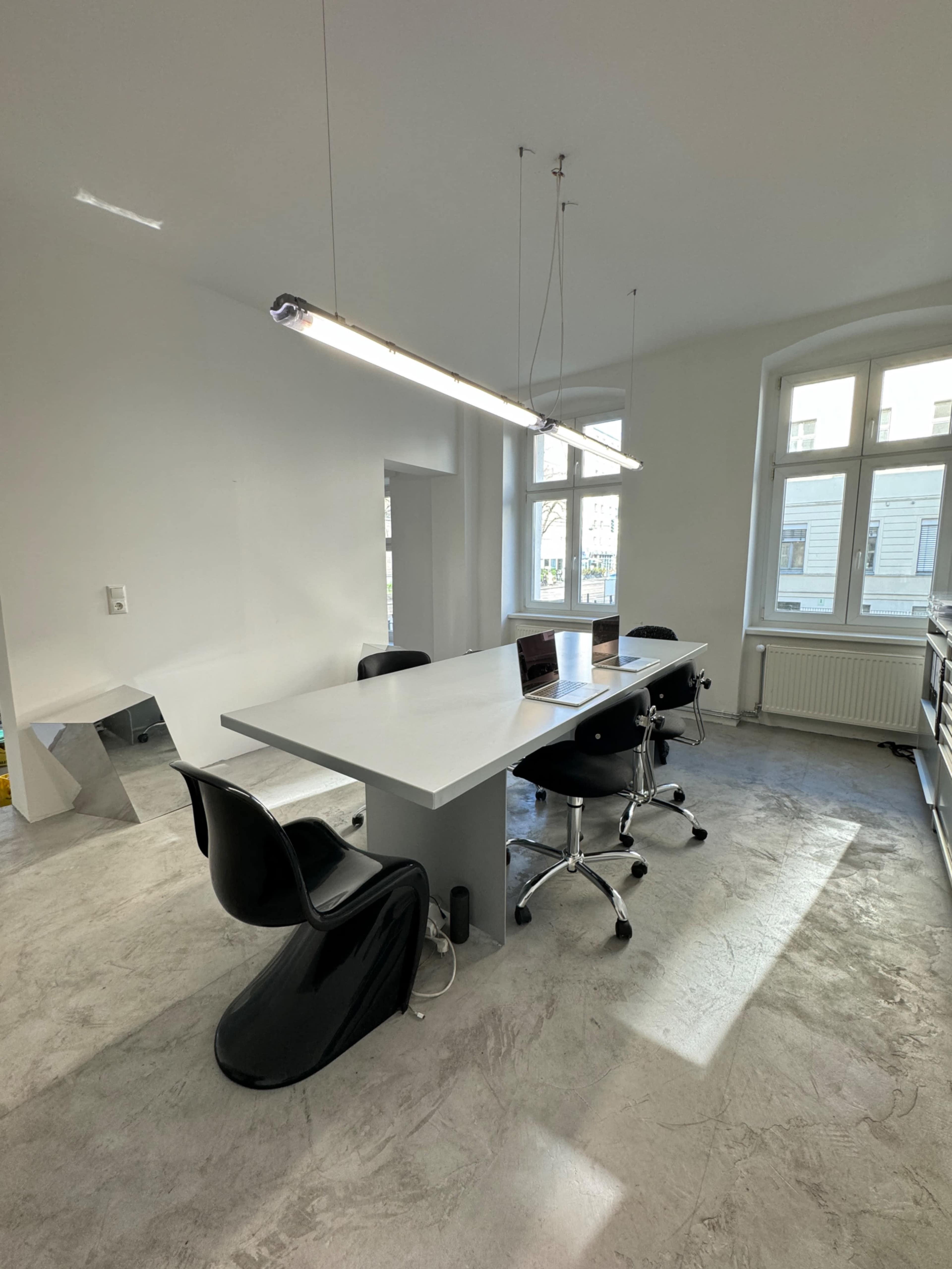 A minimalist office space features a white table surrounded by black chairs and bright overhead lighting.