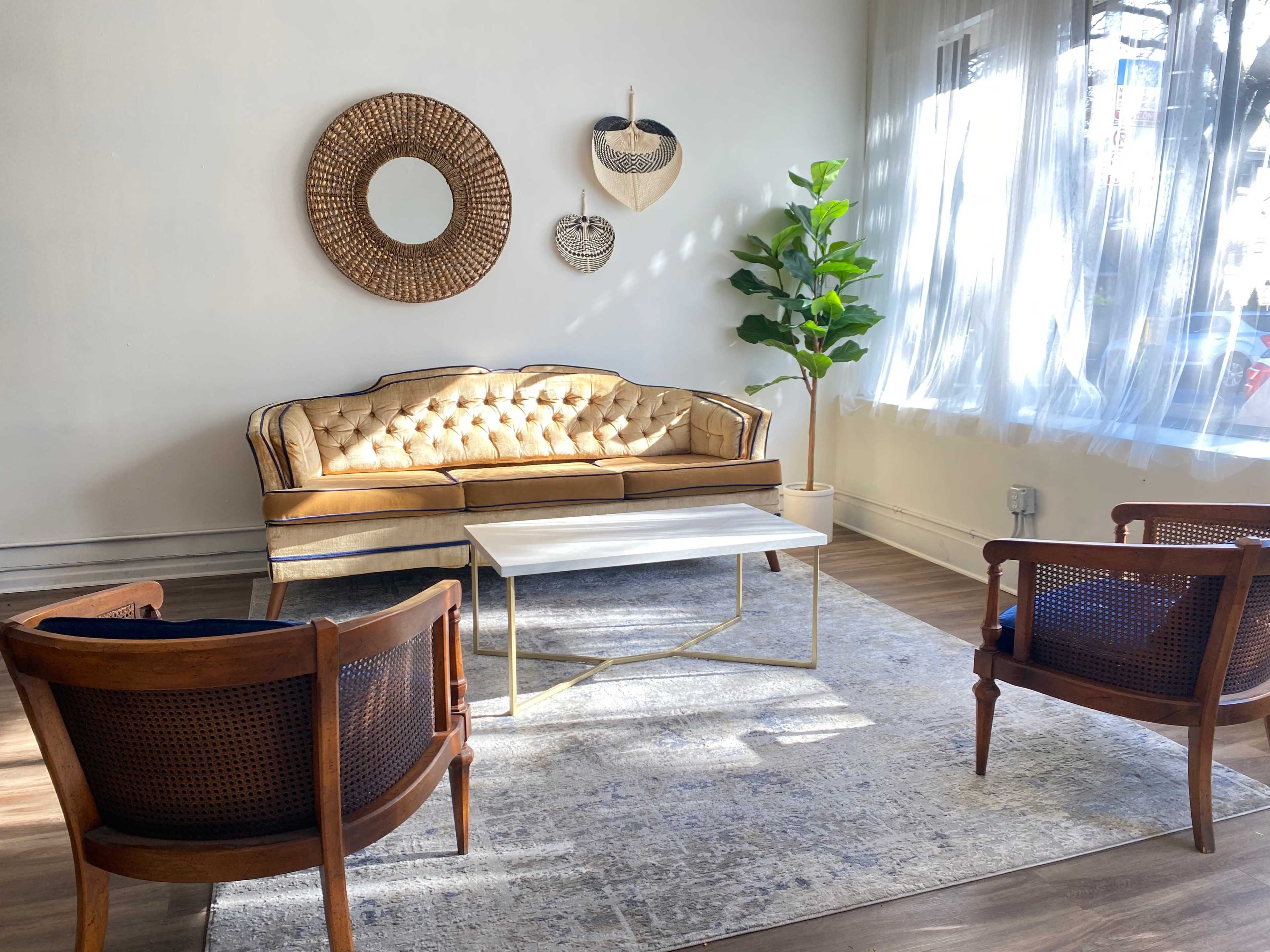 A light-colored room with a tufted beige sofa, two wooden armchairs, a coffee table, a decorative mirror, and a potted plant near a window.