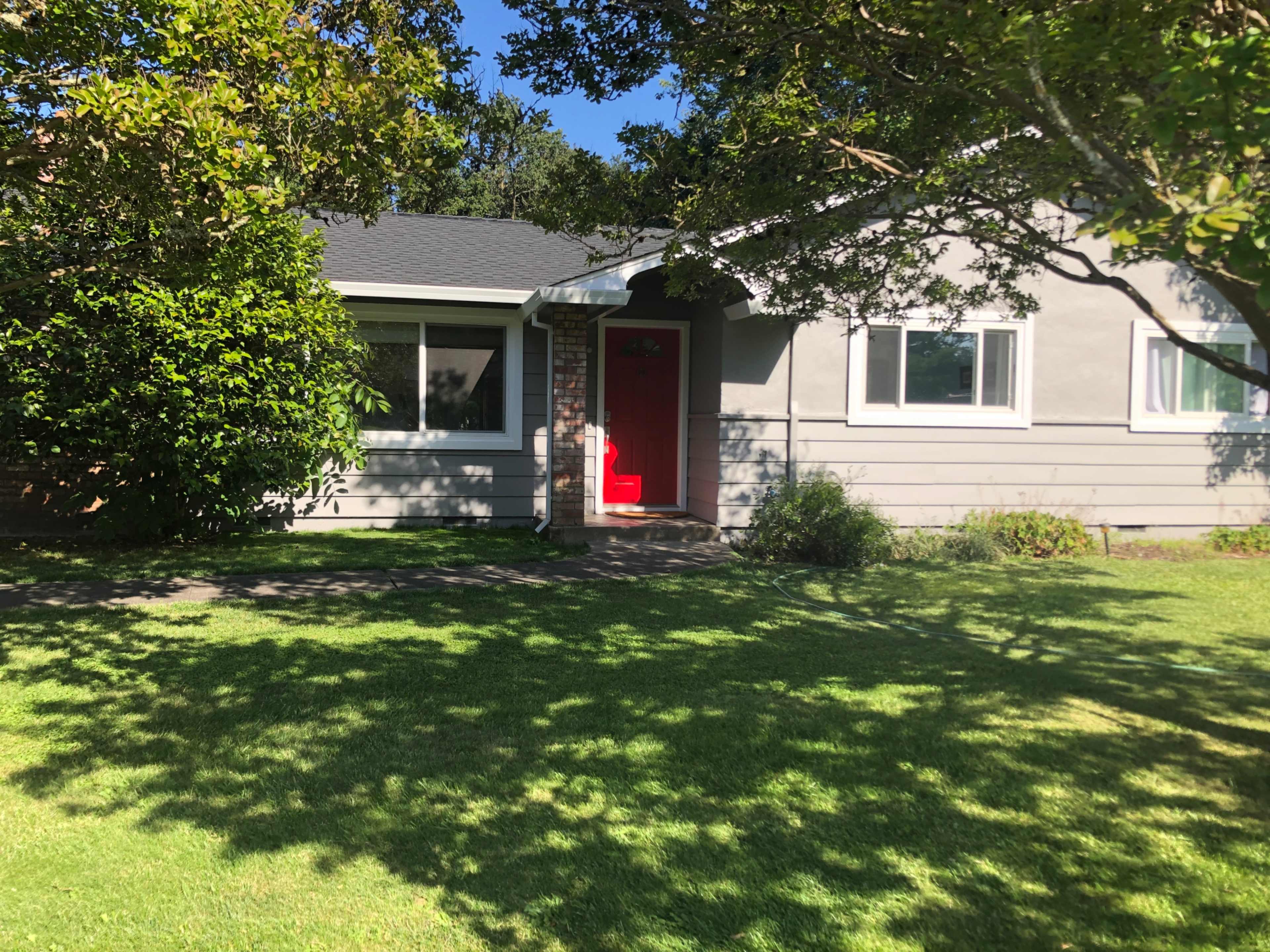 A single-story house with a red front door is set against a backdrop of green grass and trees.