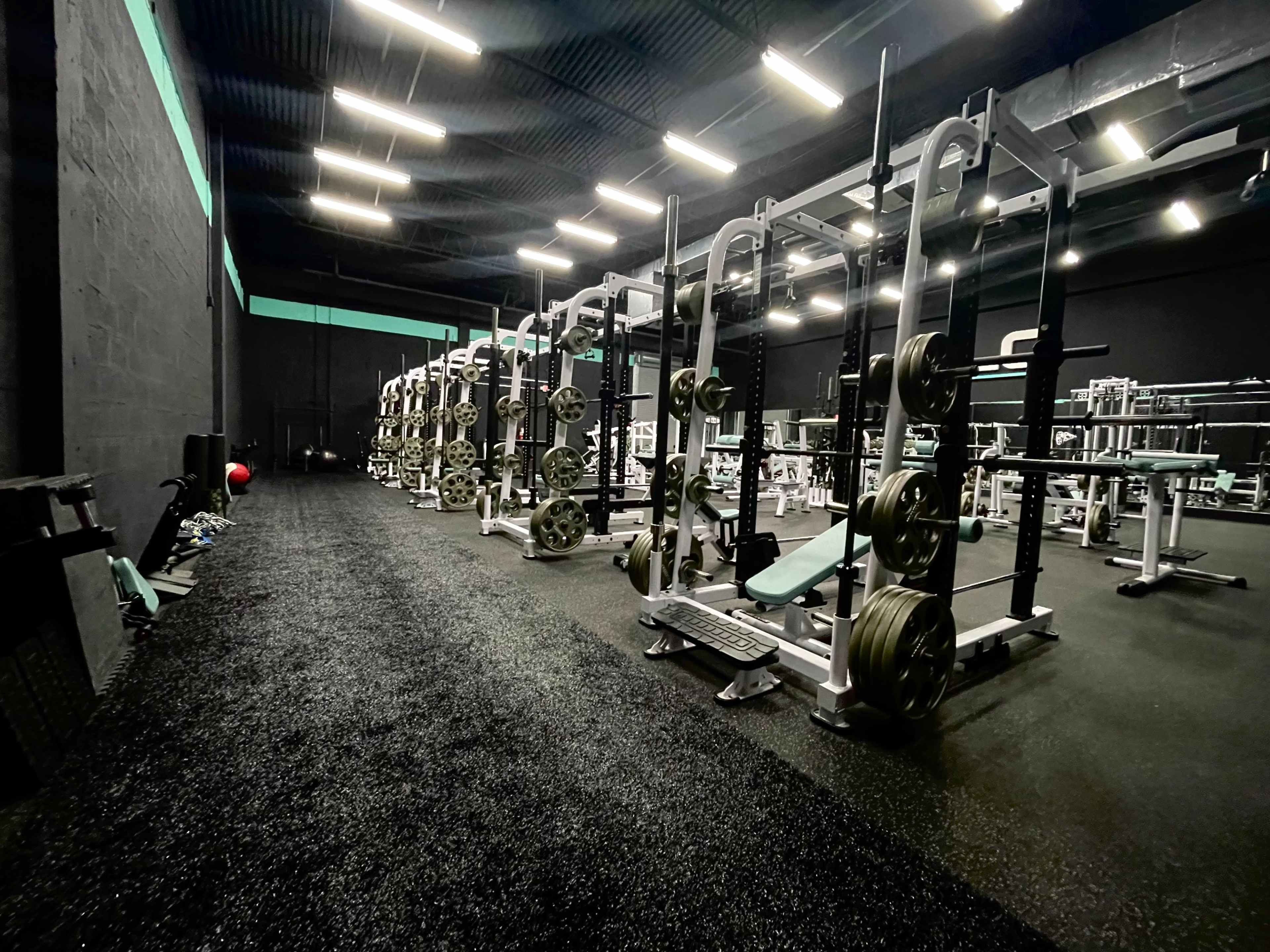 A gym interior with multiple weightlifting stations and equipment arranged along a dark, rubberized floor.