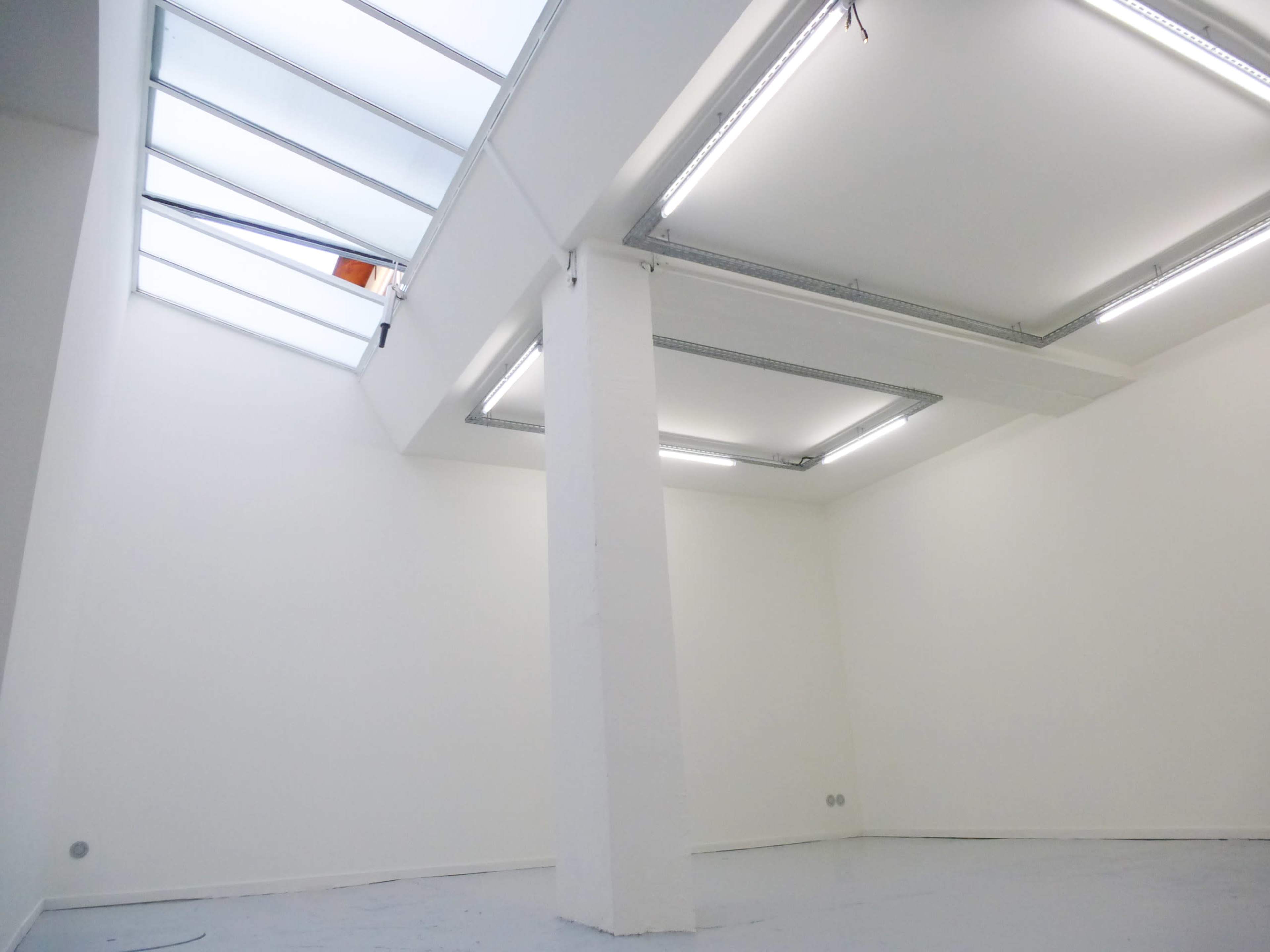 A minimalistic indoor space with white walls, a high ceiling, and skylights illuminating the room.