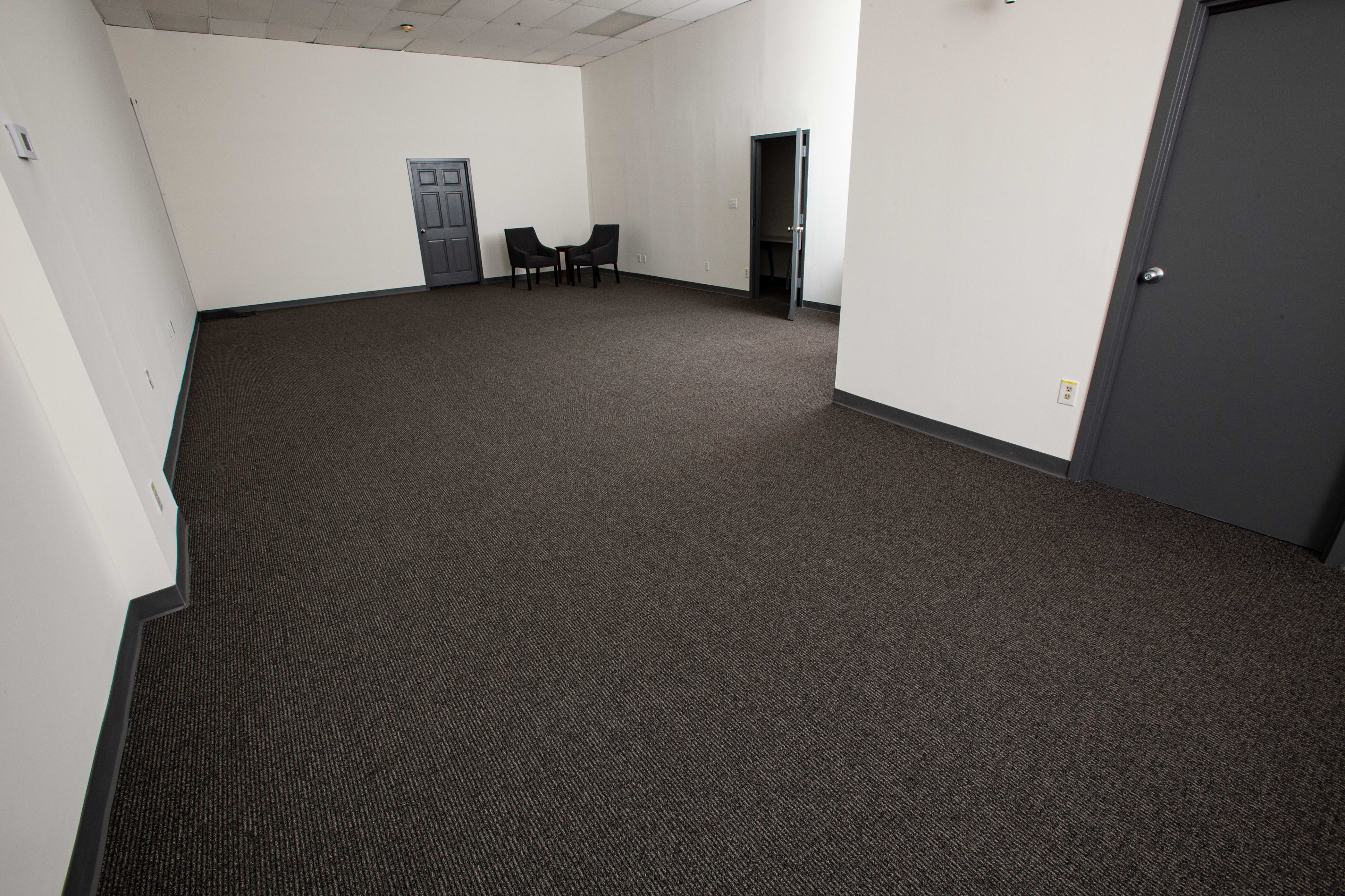 An empty room with gray walls, a dark carpeted floor, and two chairs positioned against one wall.