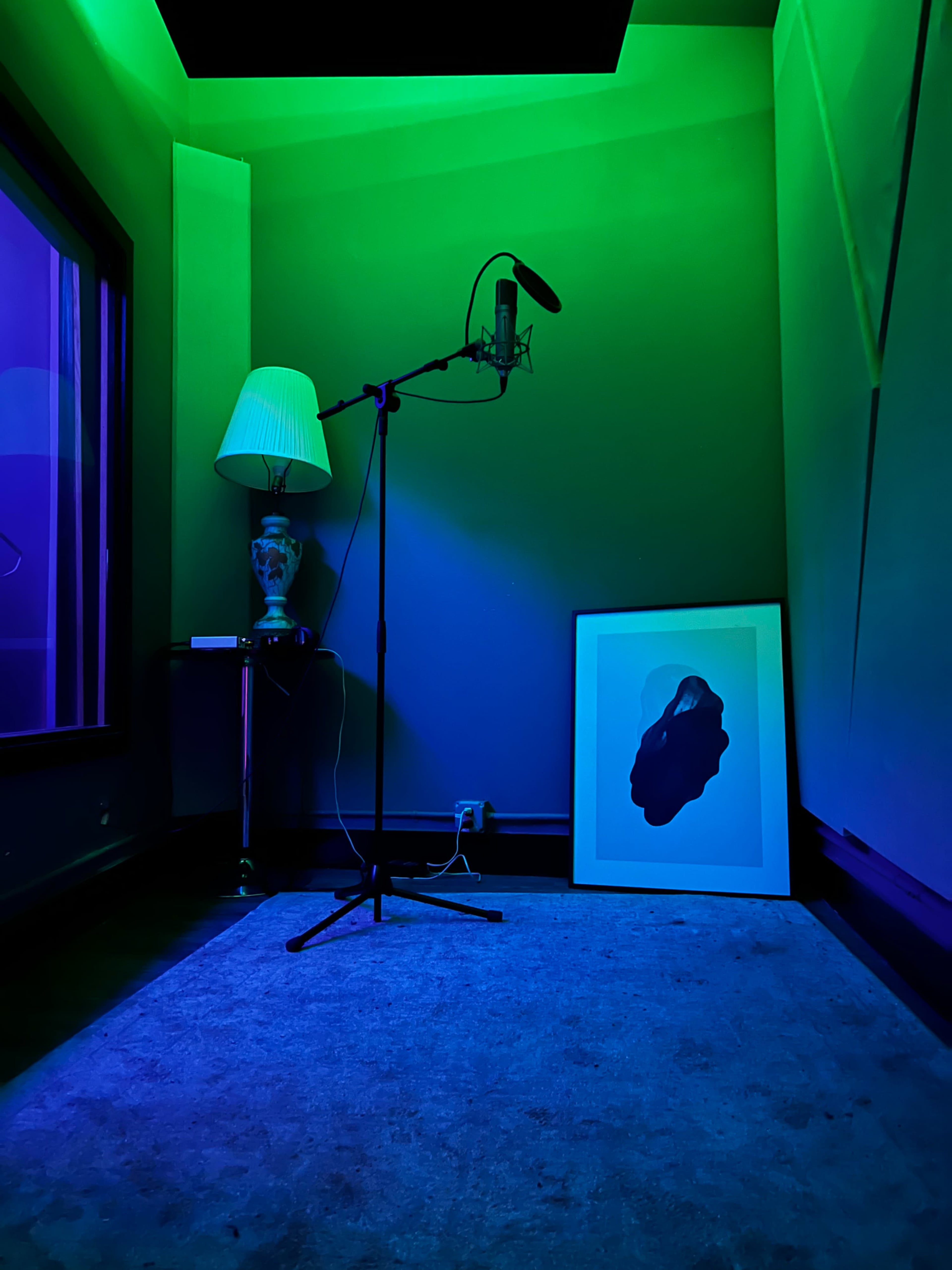 A small recording studio corner with a microphone on a stand, a lamp, and an abstract framed artwork against a wall illuminated in green and purple lights.