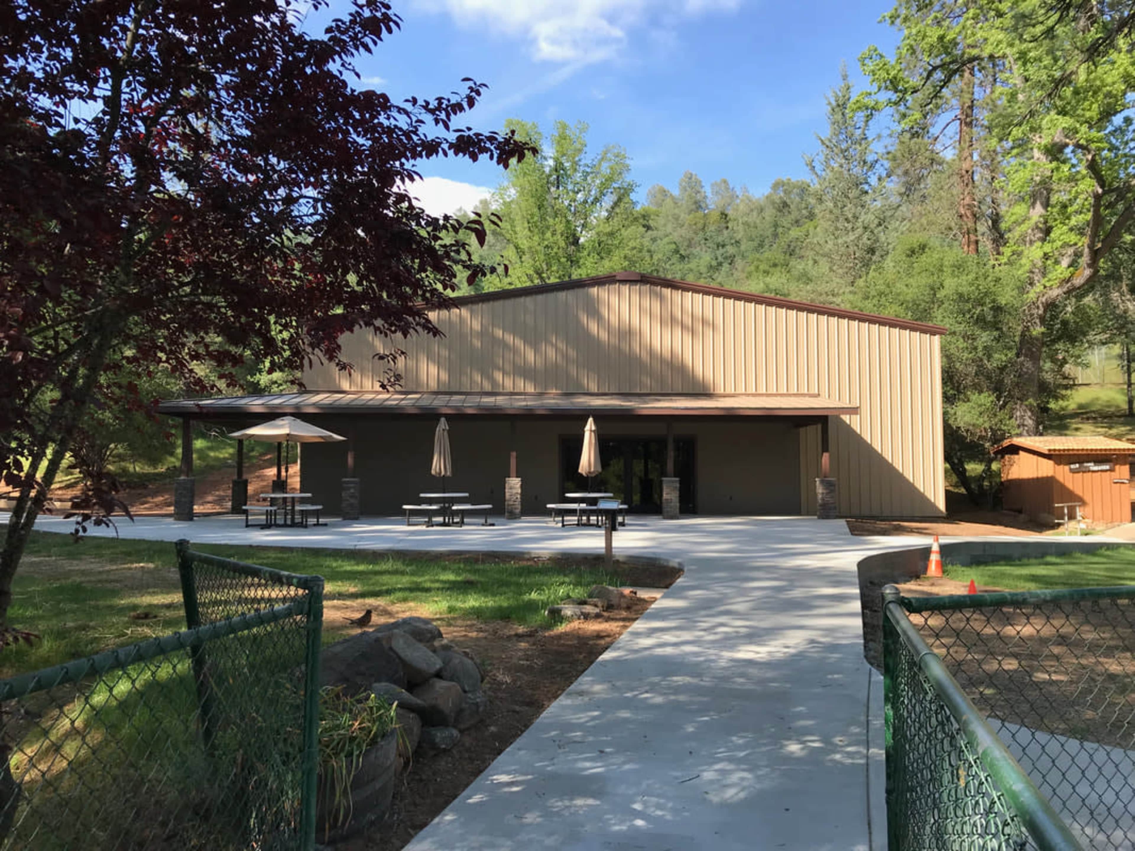 A metal-clad building with a concrete path, picnic tables, and umbrellas, surrounded by trees and green grass.