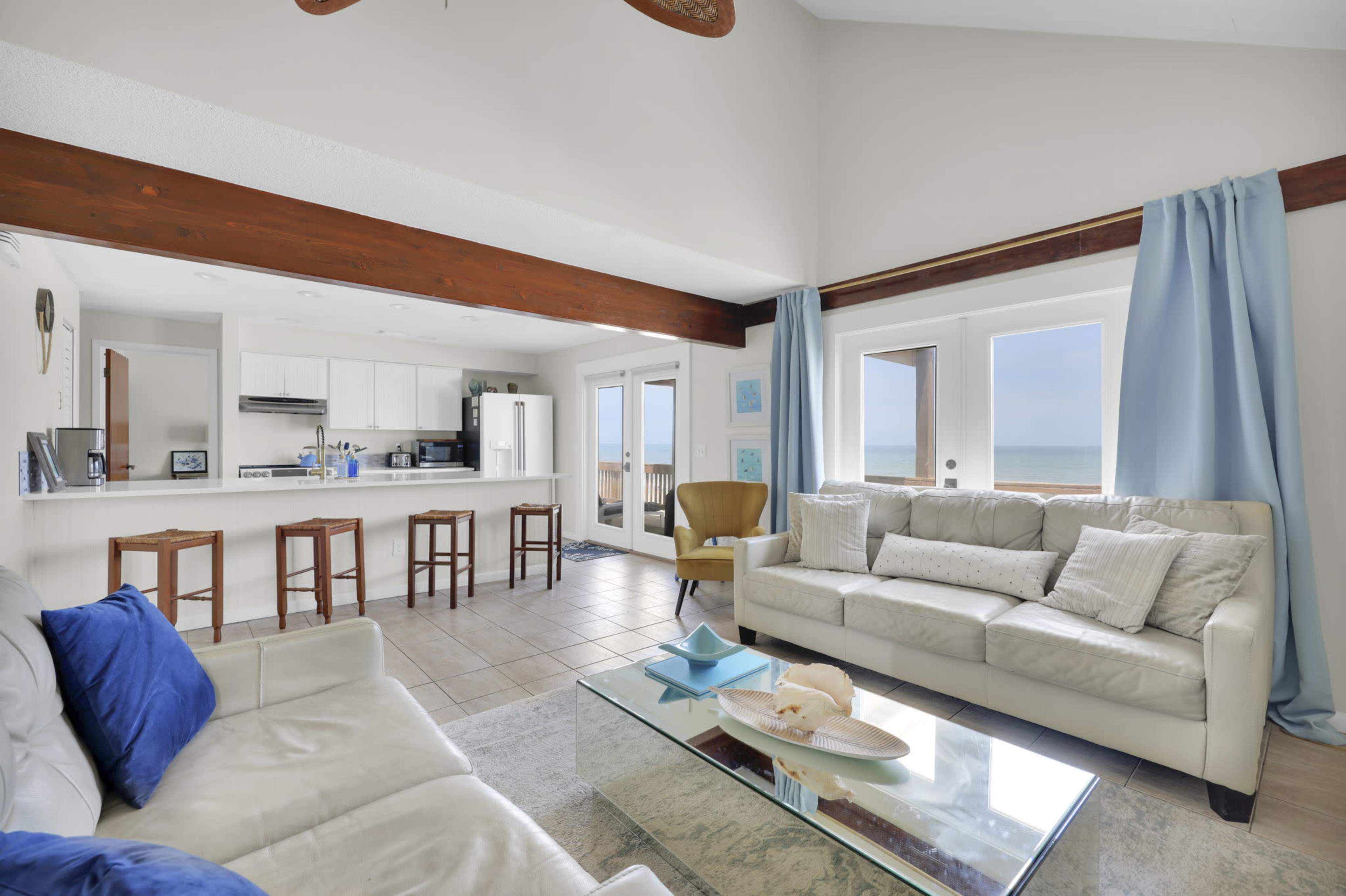 A bright, open living area features a white sofa, a glass coffee table, and large windows overlooking the ocean, with a kitchen visible in the background.
