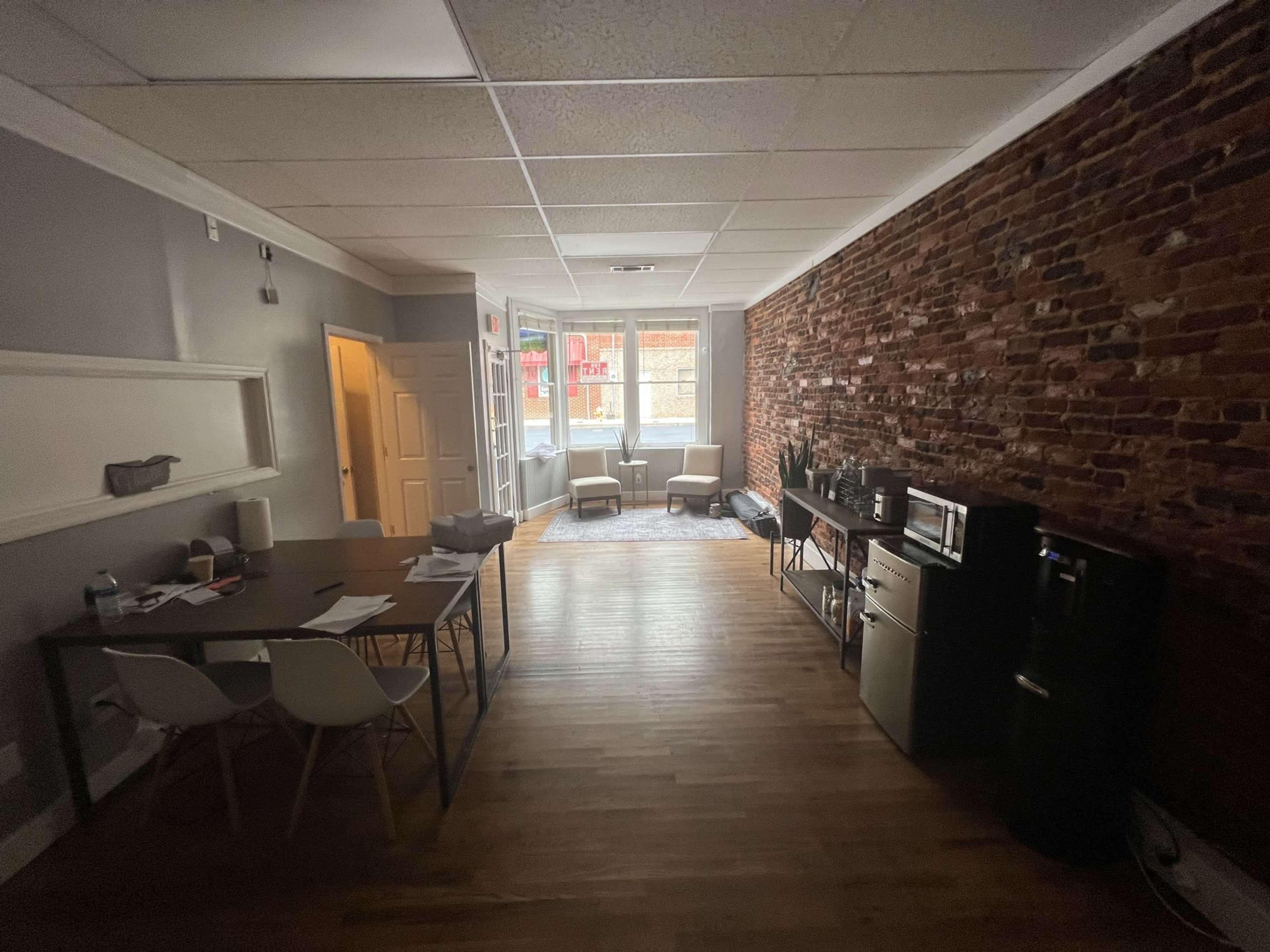 A spacious room with exposed brick walls, hardwood flooring, a dining table with chairs, and two armchairs near a window.