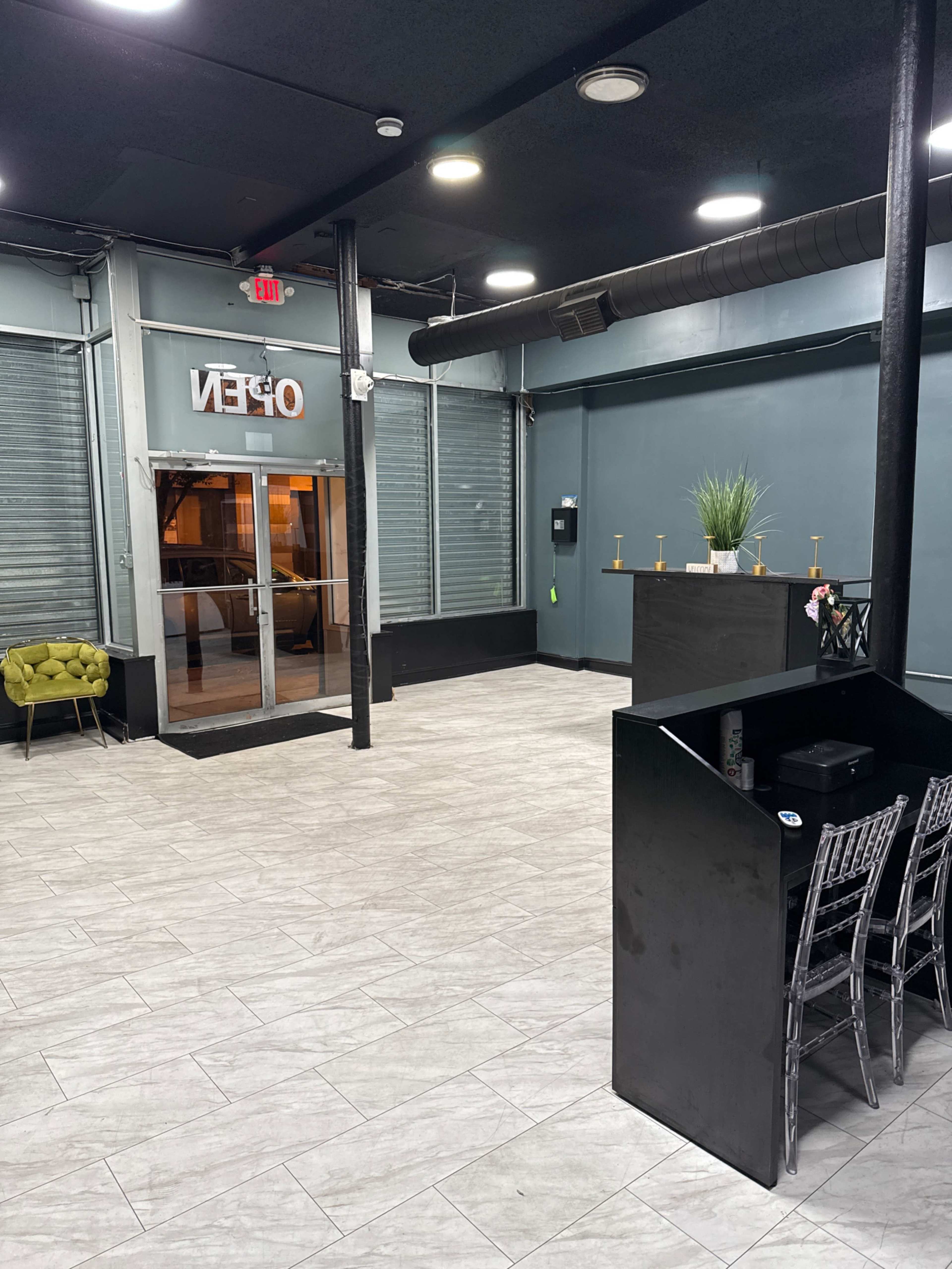 The image shows an empty interior space with gray tiled flooring, a reception area with a black counter, and large glass doors that display "OPEN."