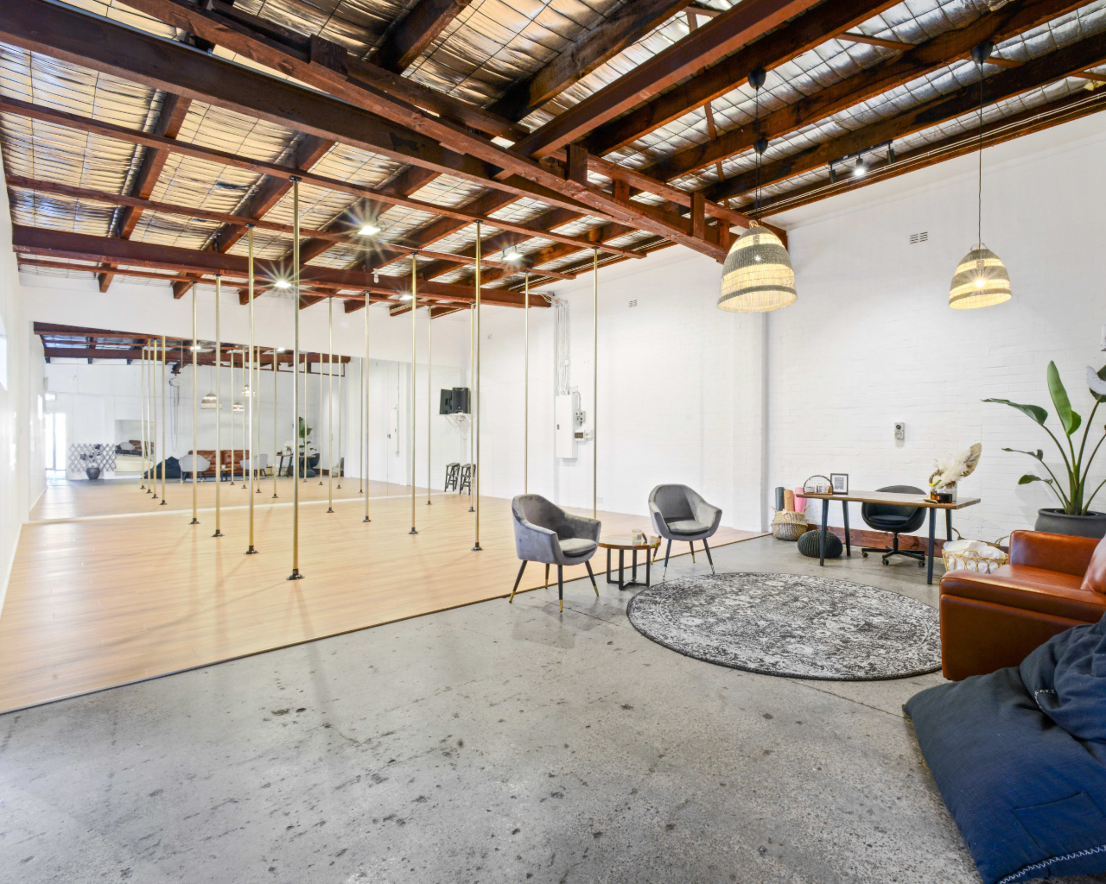 The image shows a spacious studio with wooden beams, a mirrored wall, and a mix of seating areas, including chairs and rugs, on a polished concrete floor.