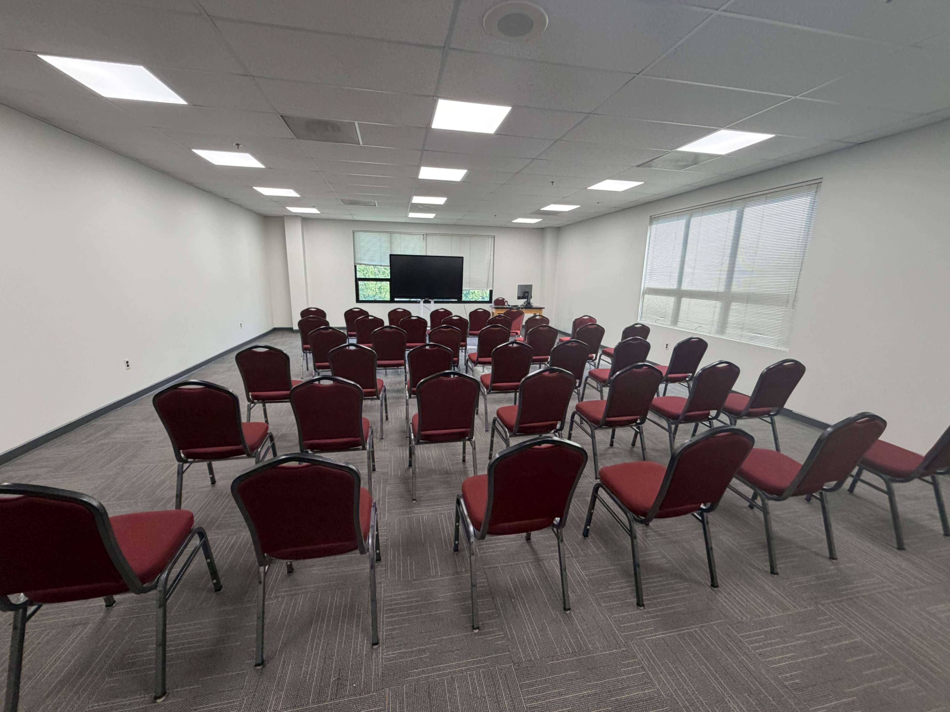 The image shows a meeting room with a set of red chairs arranged in rows facing a central screen.