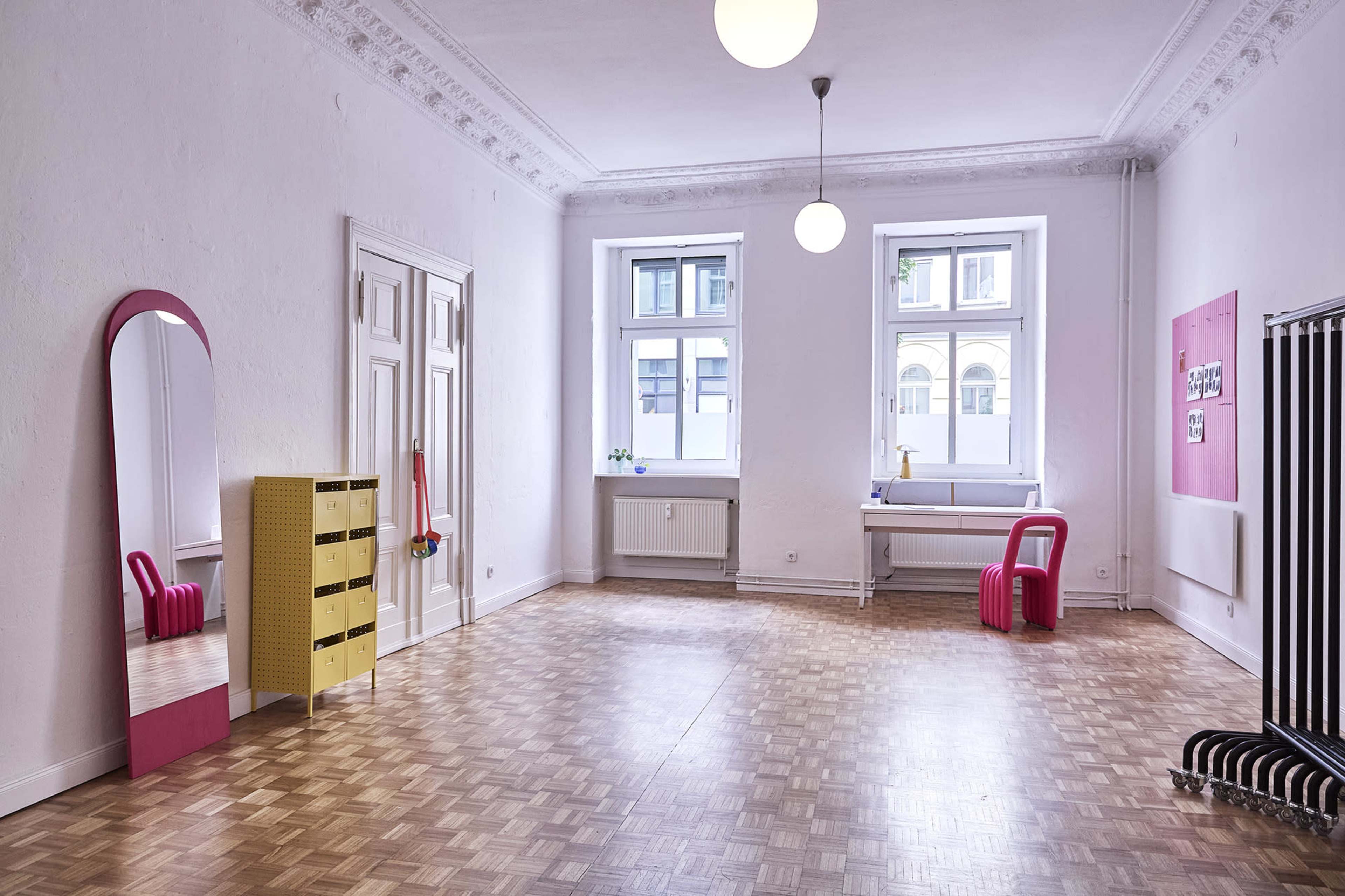 The image shows a spacious, minimalist room with light-colored walls, large windows, a pink mirror, a yellow storage unit, and wooden flooring.