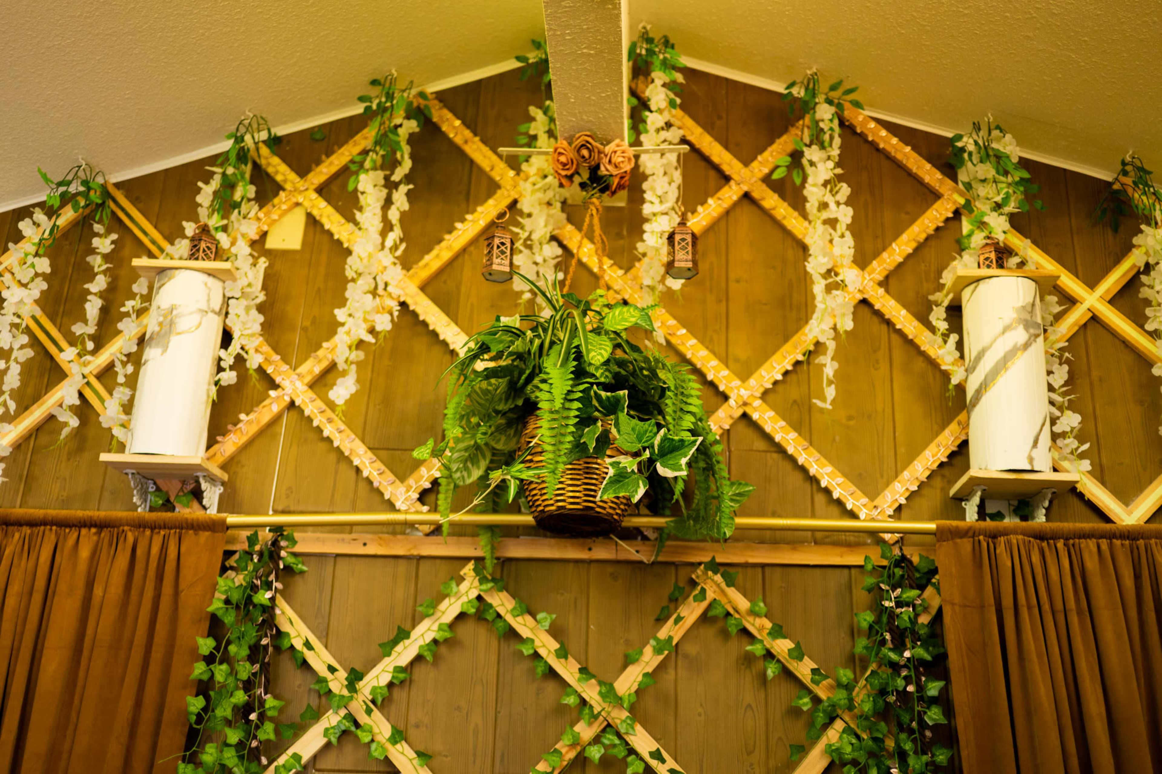The image shows a wooden wall adorned with decorative trellis patterns and hanging plants, complemented by white floral arrangements and lanterns.