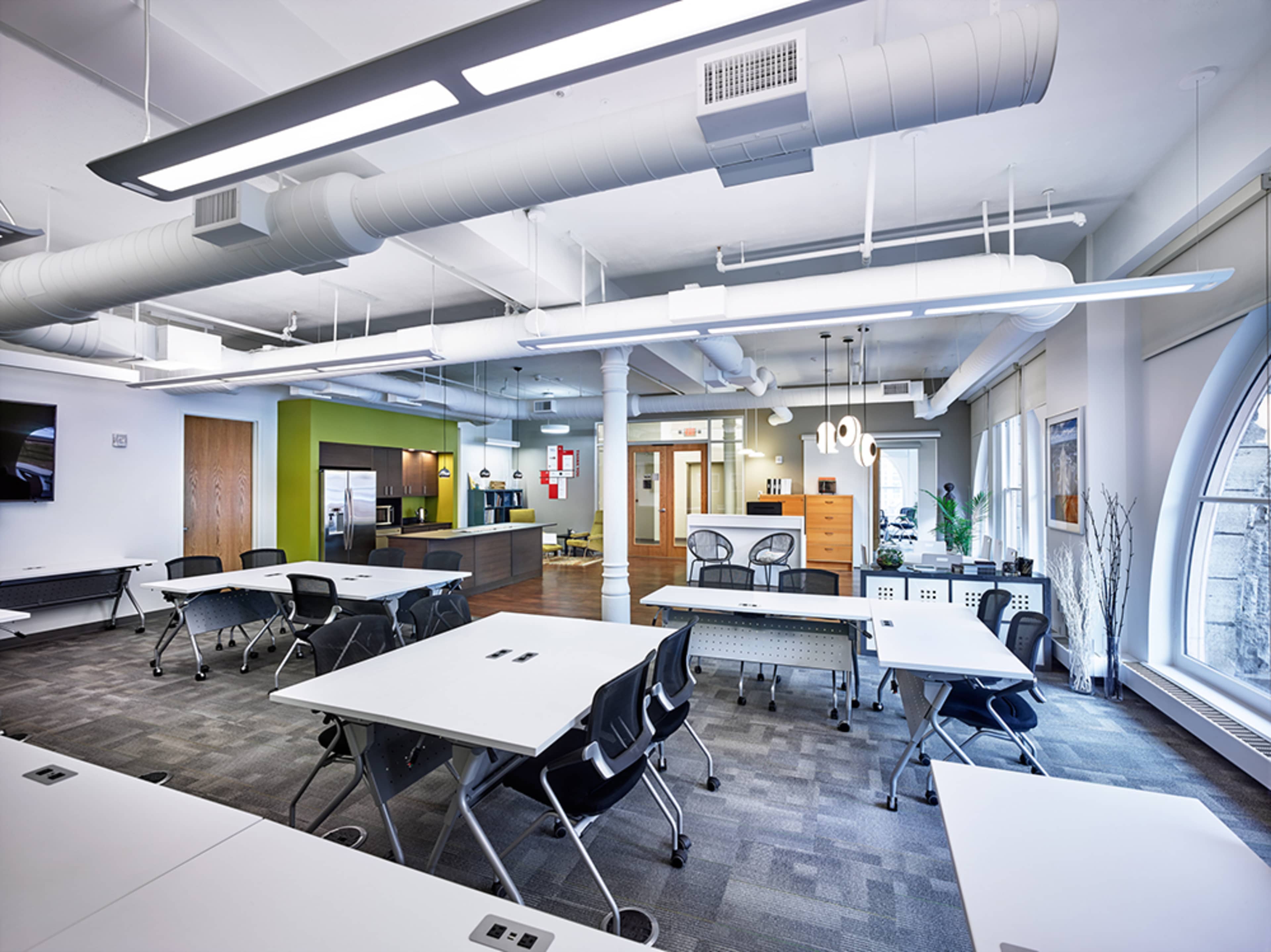 A modern classroom or meeting space with modular tables, ergonomic chairs, and a kitchenette area in the background.