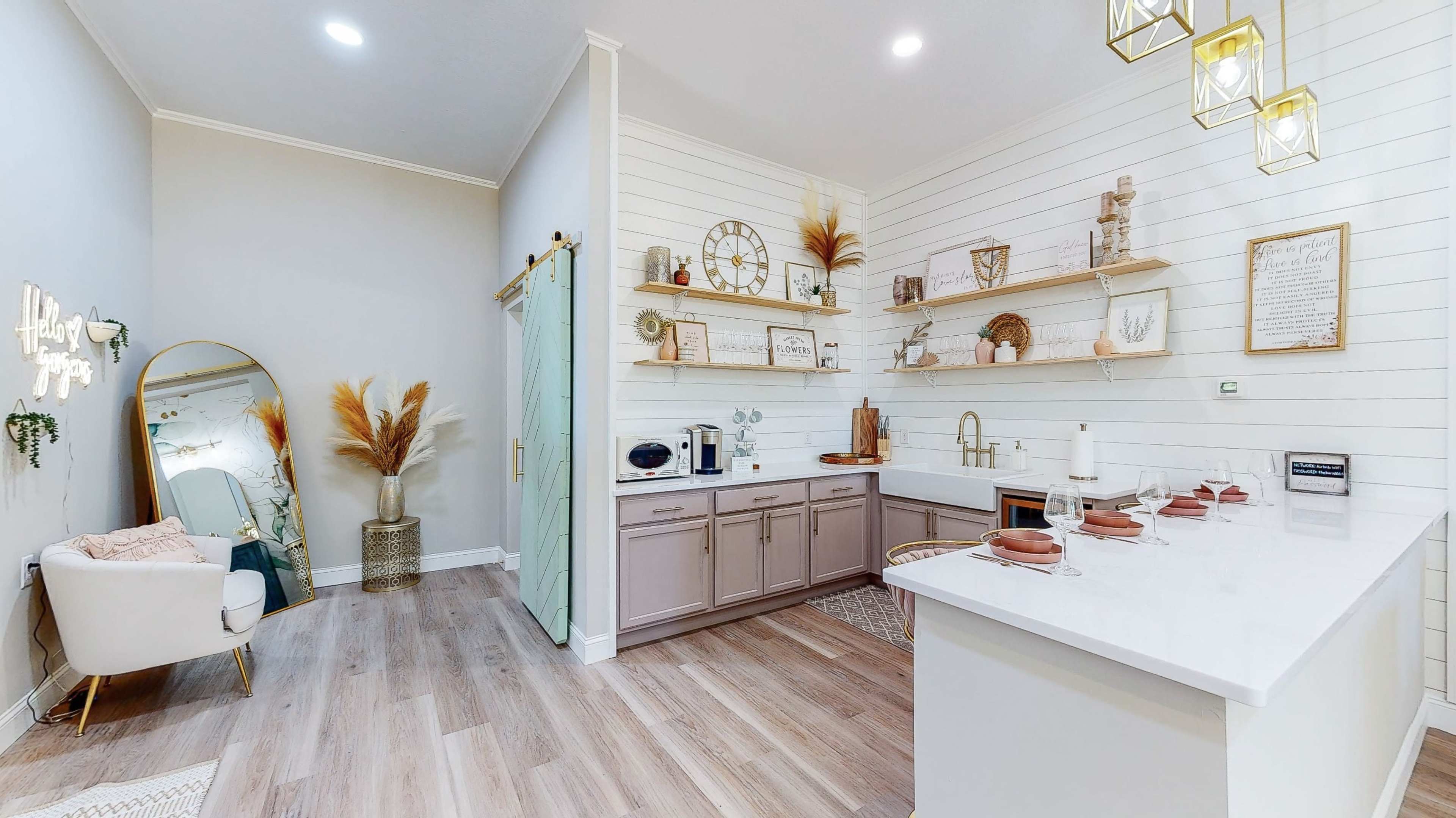 A modern kitchen and living area featuring a white wall with decorative shelves, a pastel-colored kitchen cabinet, and a seating area with a mirror and decorative plants.