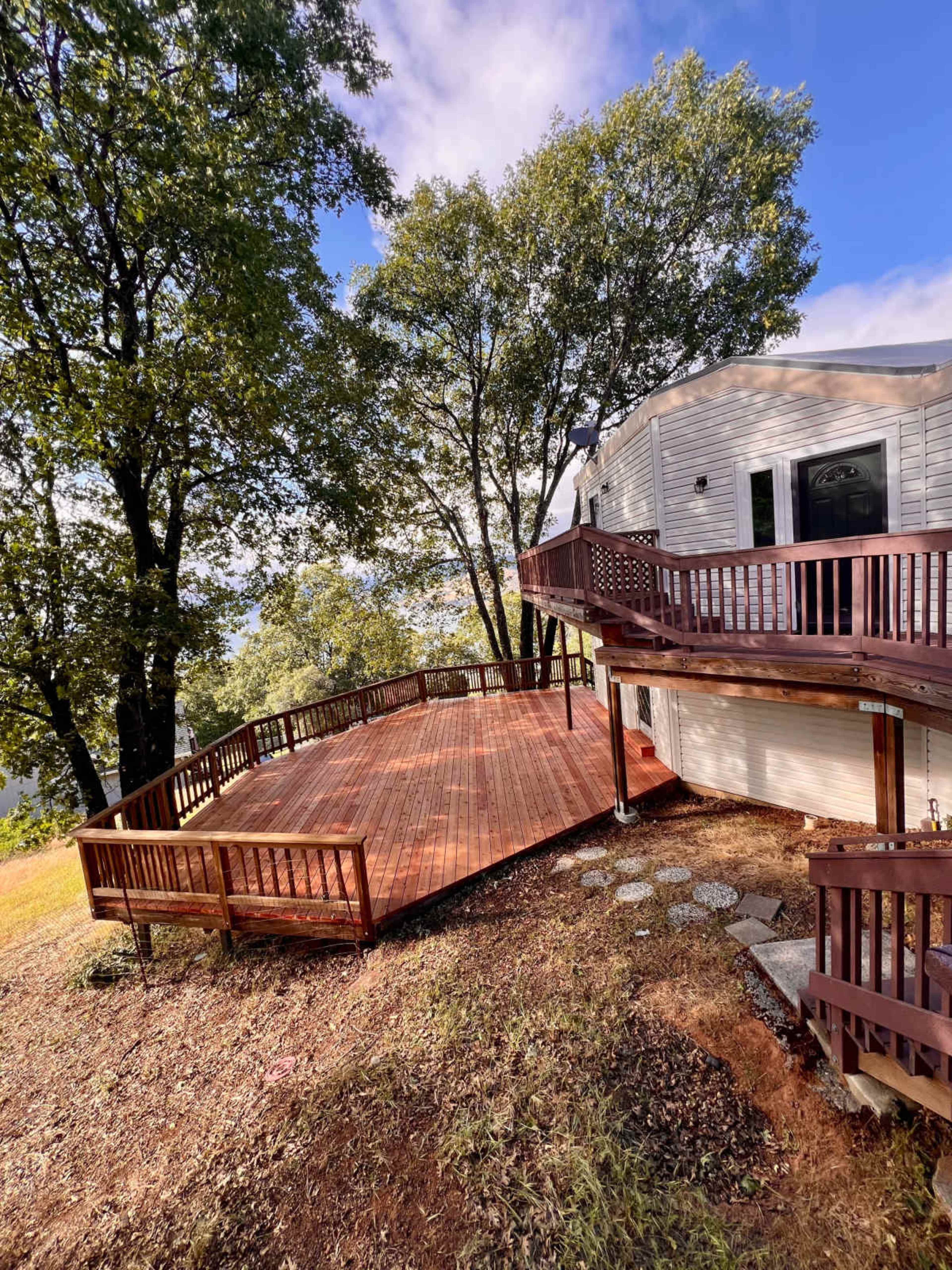 The image shows a large wooden deck extending from a white house, surrounded by trees and a sloped yard.