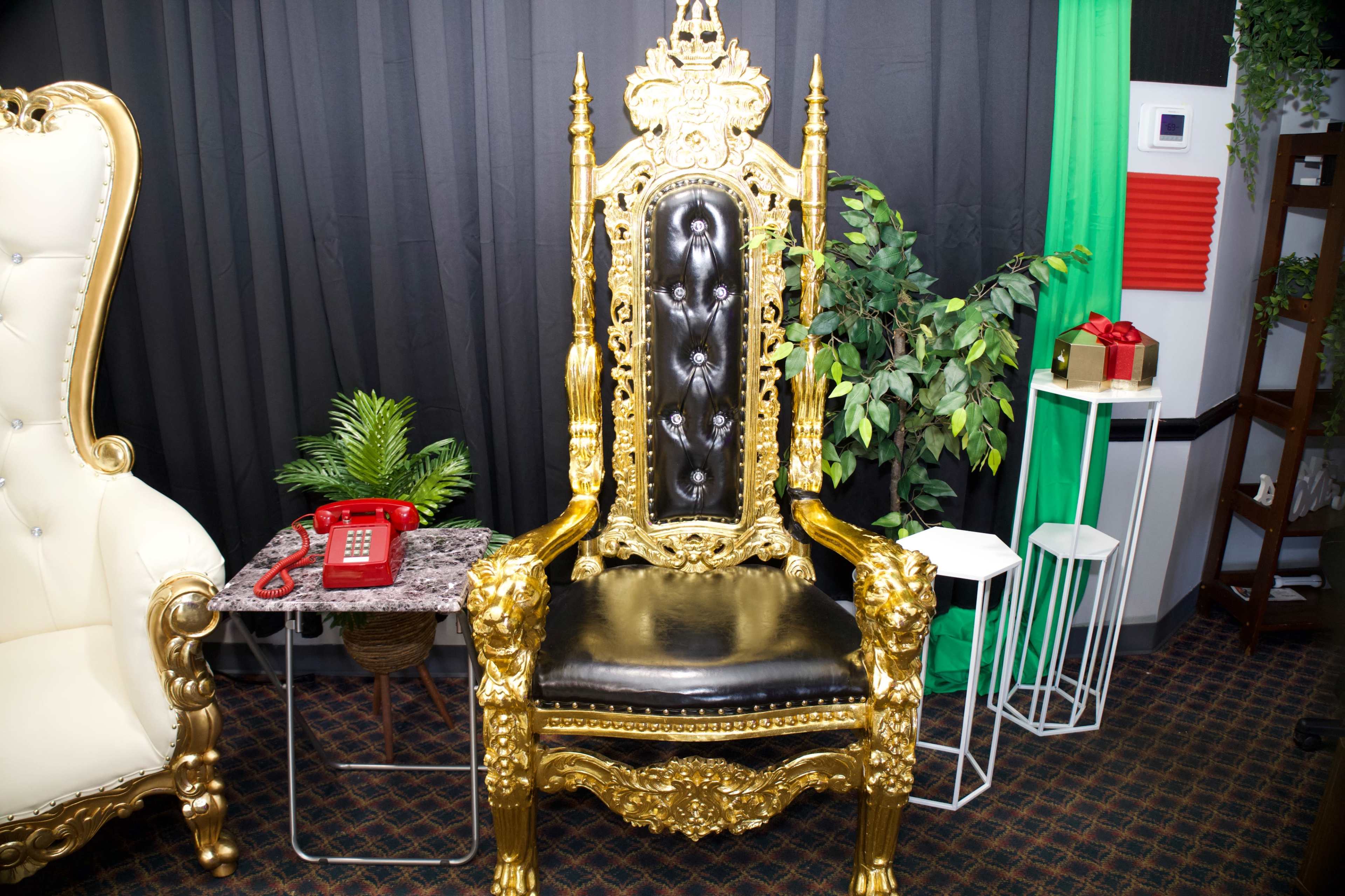 An ornate gold throne with black upholstery positioned next to a red retro telephone and potted plants against a black backdrop.