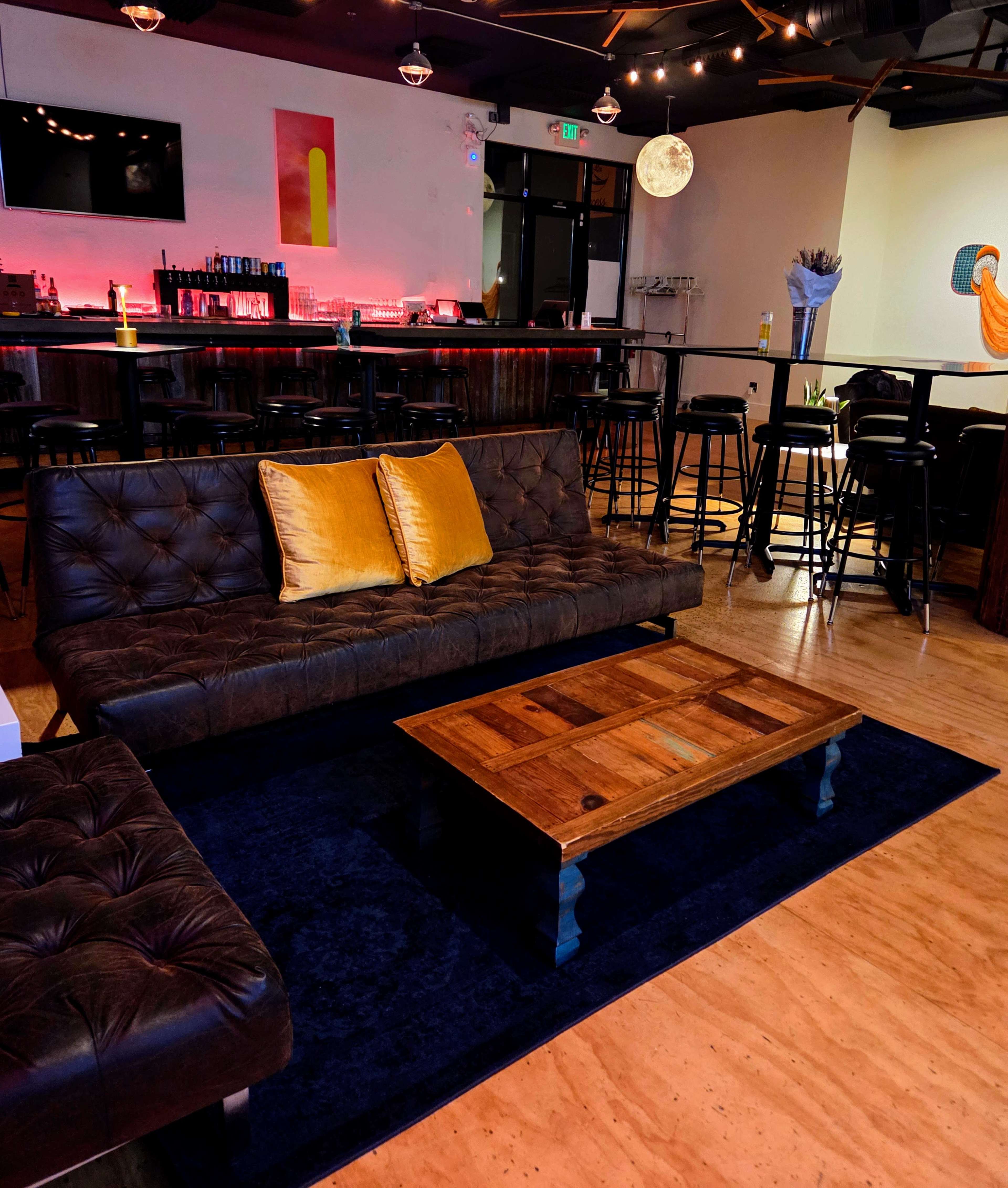 The image shows a cozy lounge area with a dark leather couch, a wooden coffee table, and a bar in the background illuminated by soft lighting.