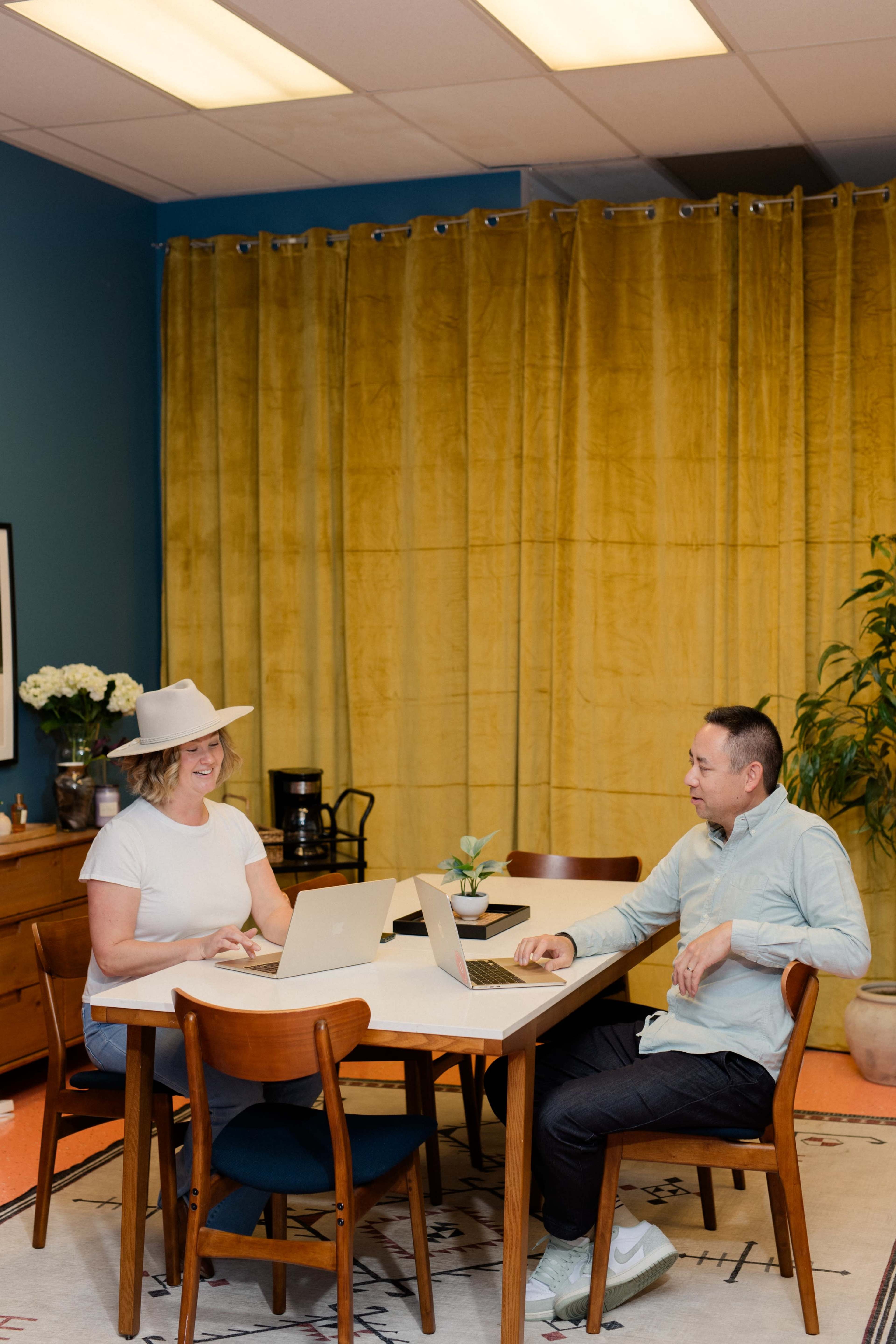 Two people are sitting at a table with laptops in a room featuring blue walls and yellow curtains.