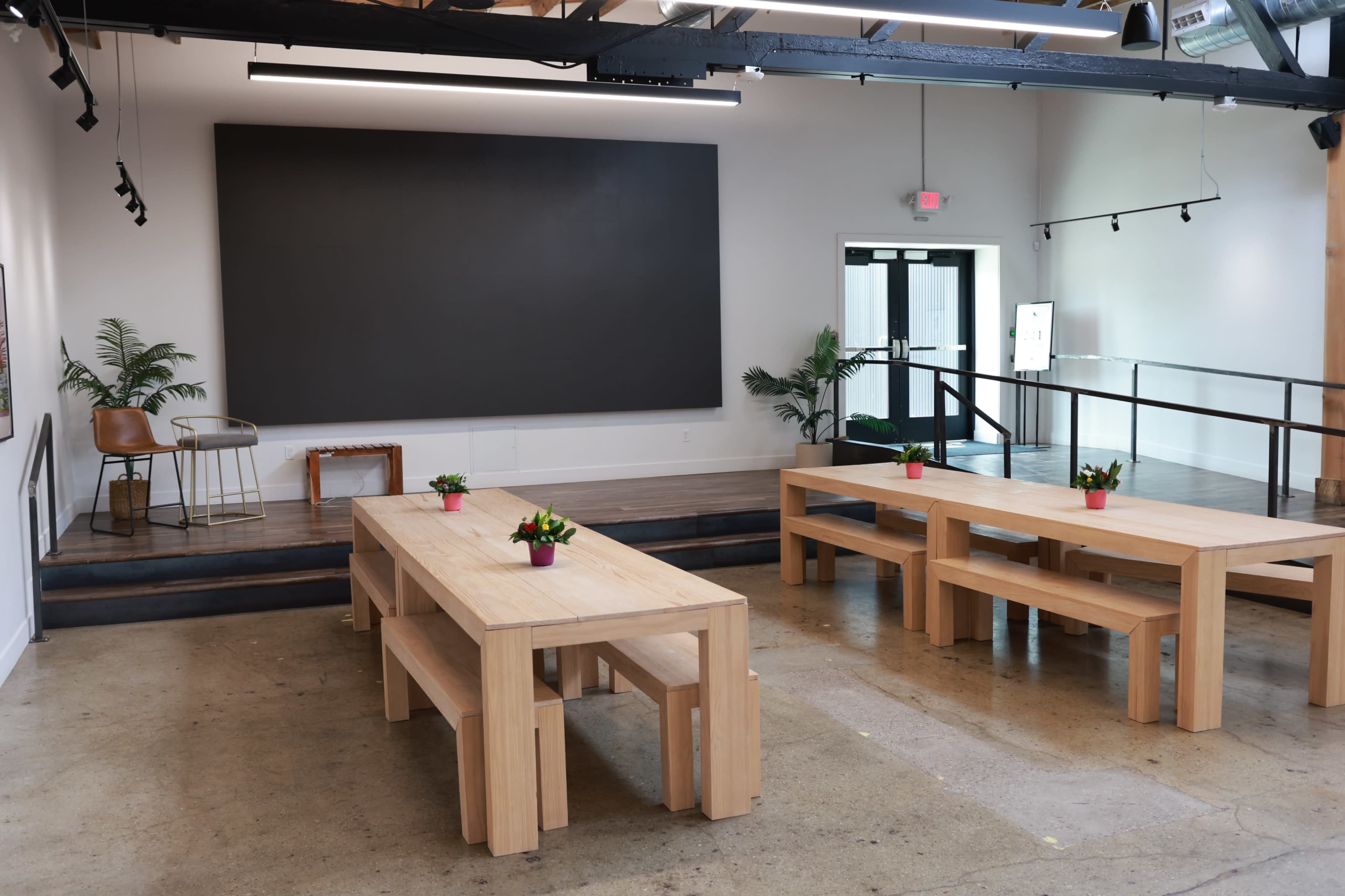 The image shows a modern multipurpose room with two large wooden tables, potted plants on each table, and a stage area featuring a black wall and a seating arrangement.