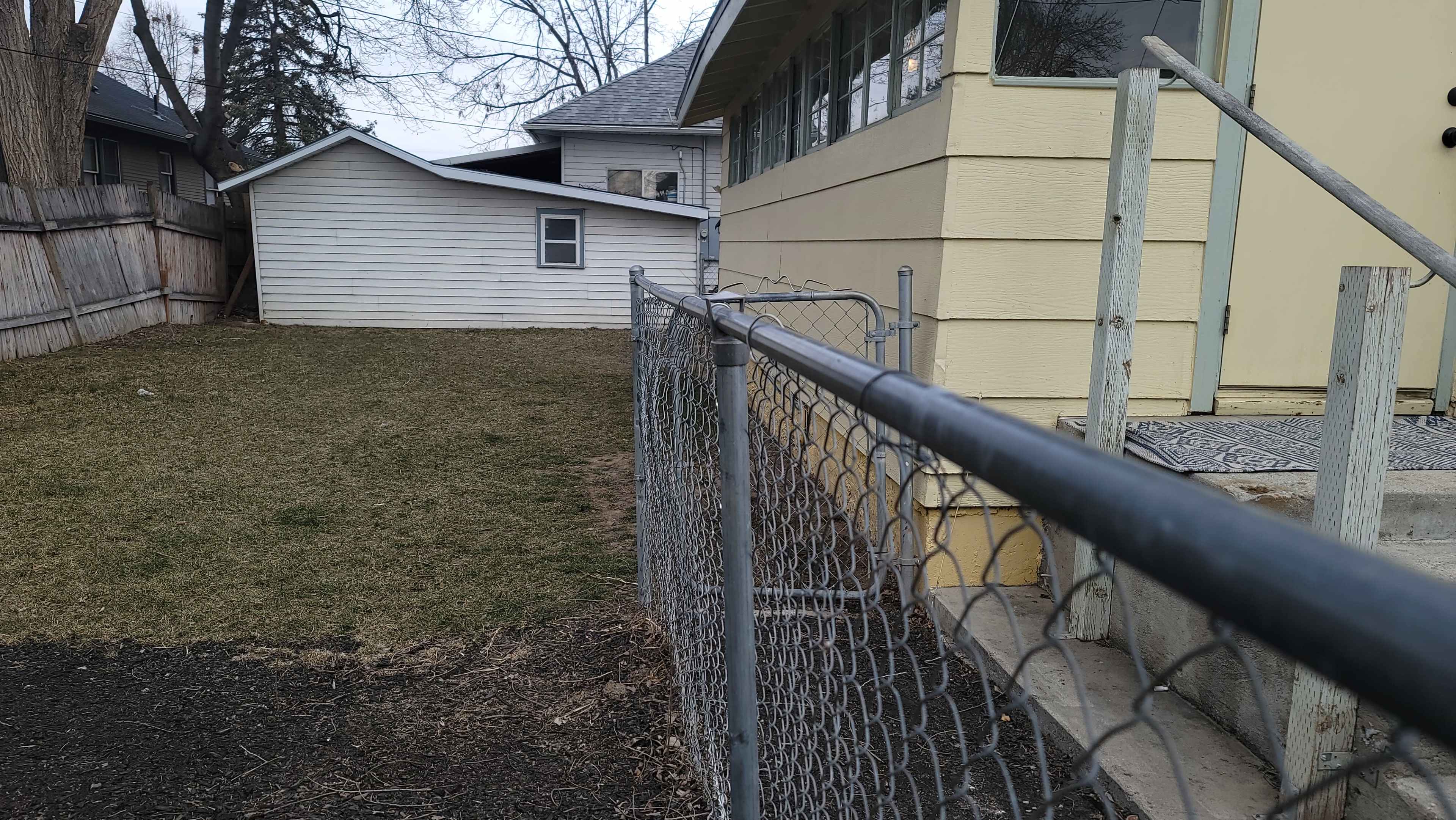 A chain-link fence runs along the edge of a grassy yard next to a small white shed and a house with a large window.