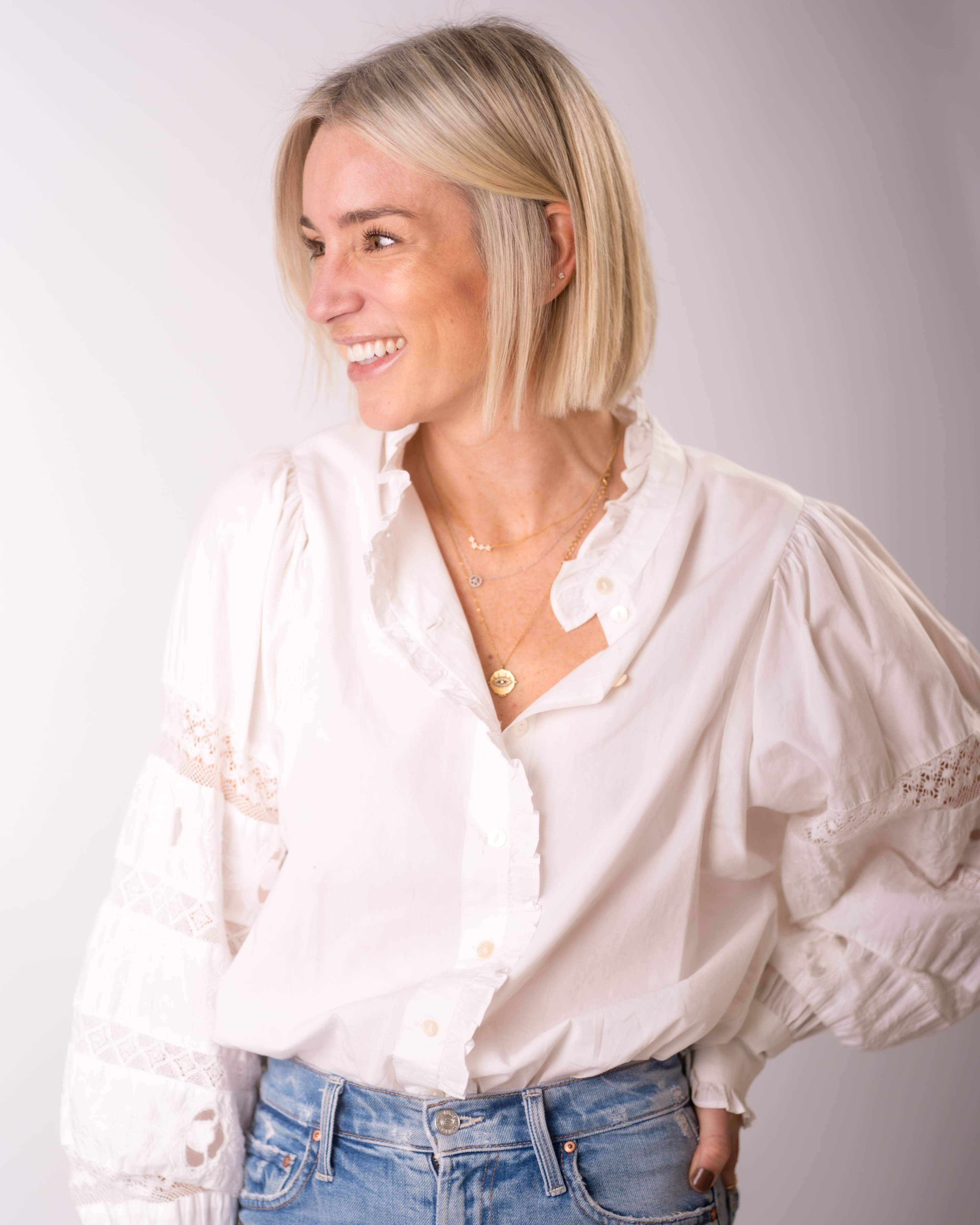 A woman with short blonde hair smiles while wearing a white blouse and blue denim jeans against a neutral background.