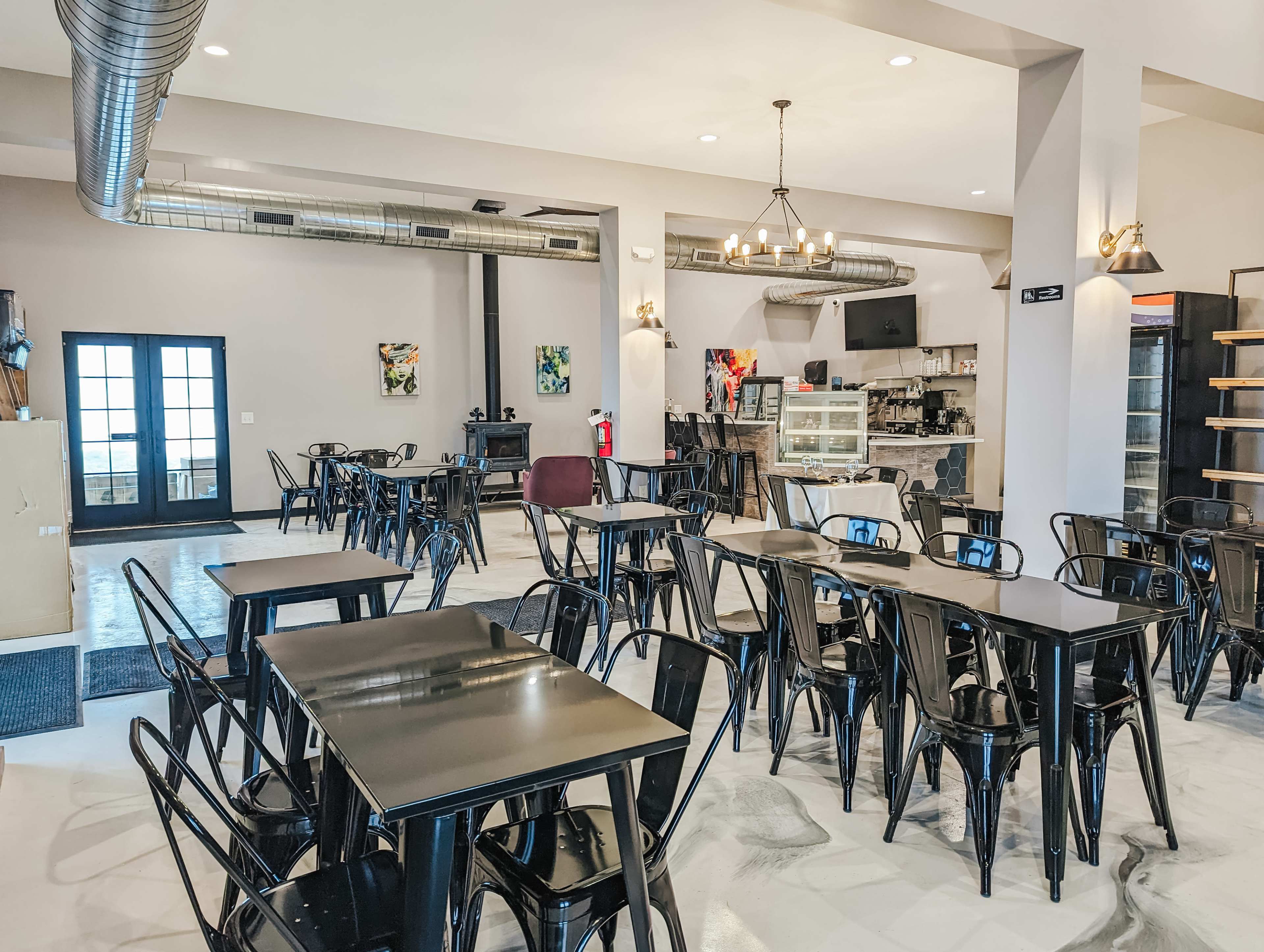 A modern café interior with black metal tables and chairs arranged on a marble floor, featuring large windows and a counter with display cases at the back.