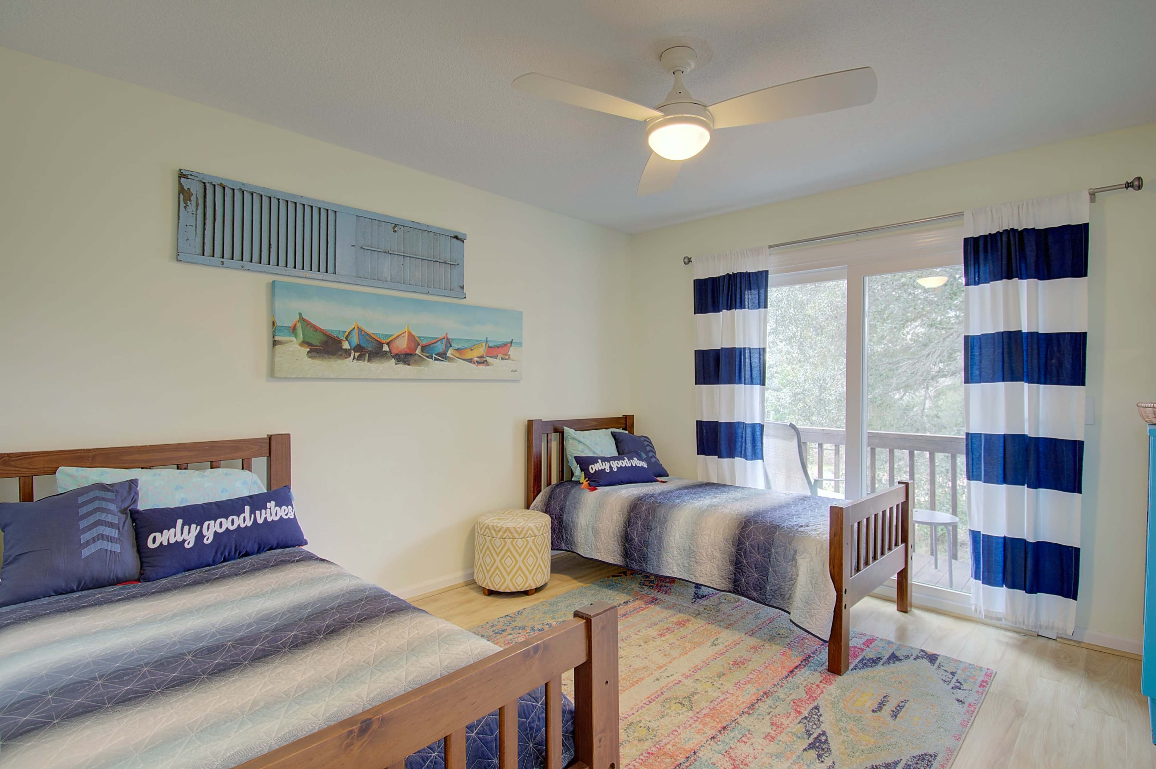 A bright bedroom with two wooden twin beds, a colorful rug, a ceiling fan, and large windows dressed with striped curtains.