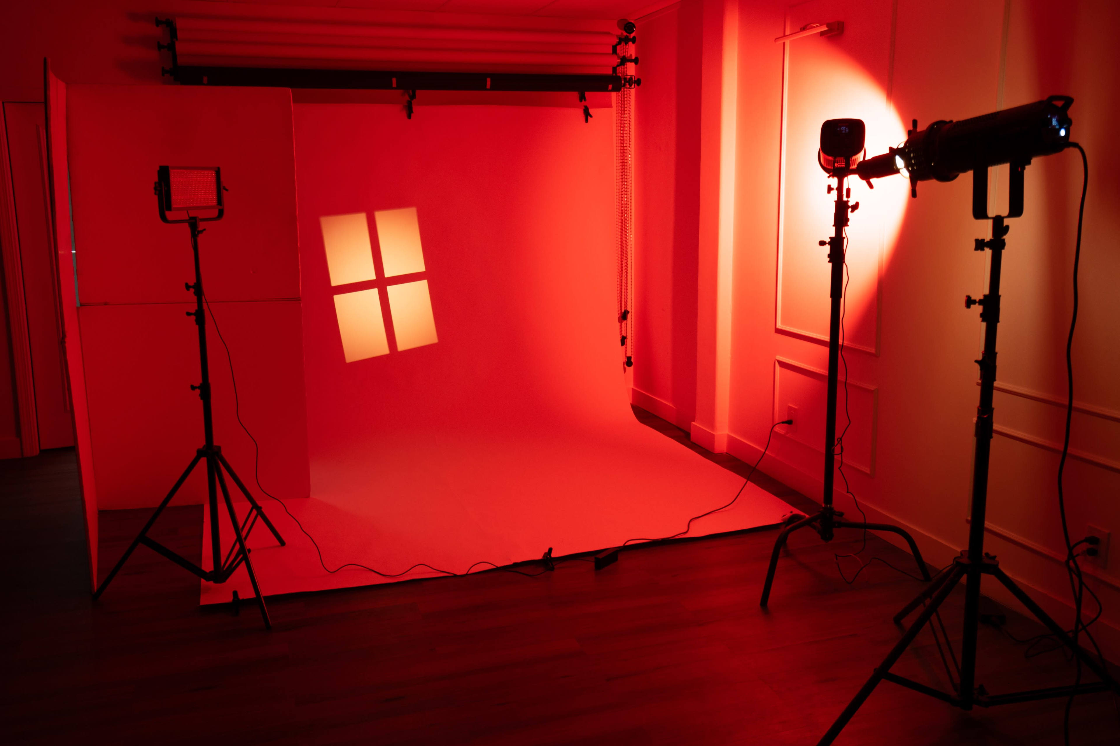 A photography studio is set up with a red backdrop and shadow patterns resembling a window on the wall, illuminated by two studio lights.