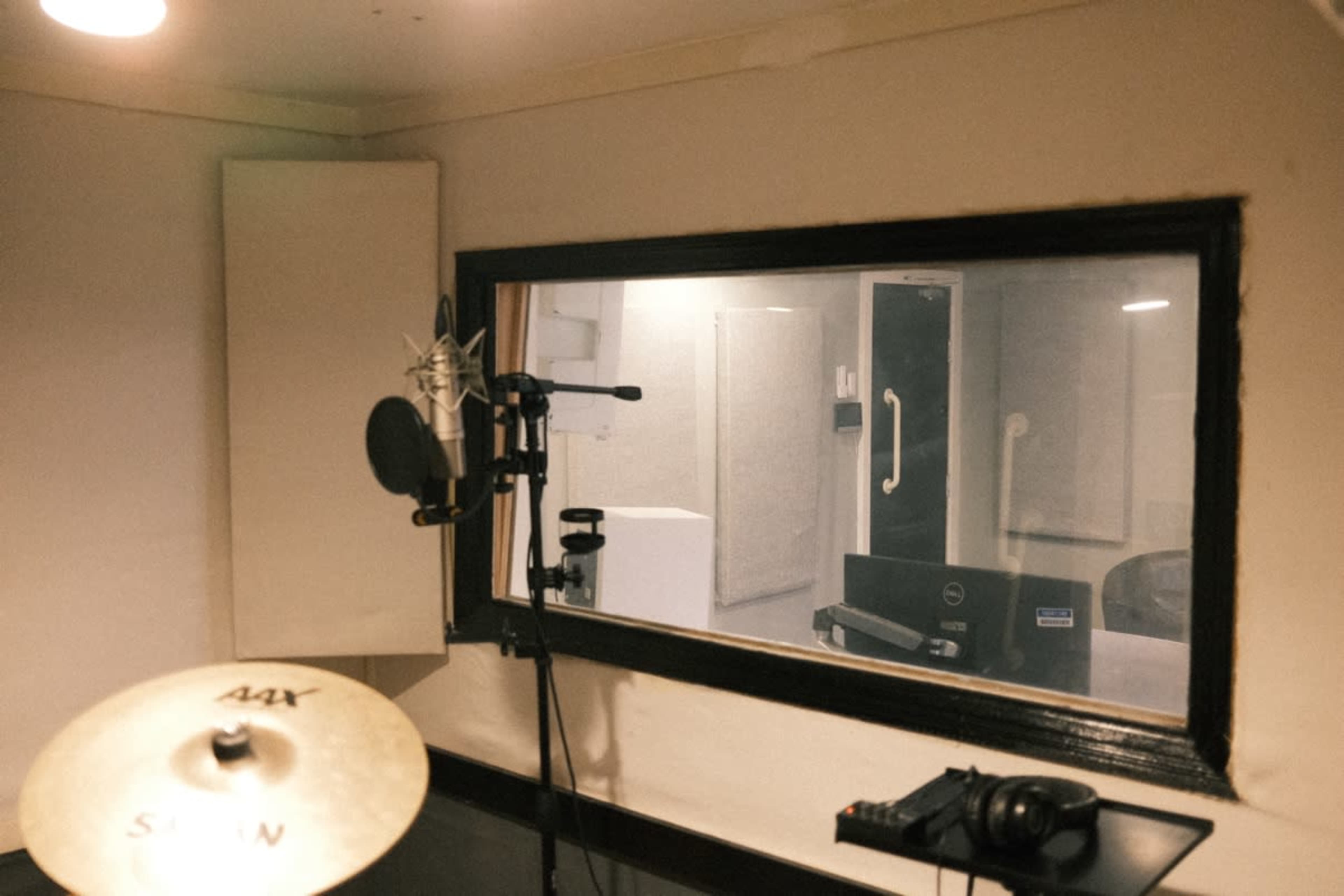 The image shows a recording studio with a microphone and a drum cymbal in the foreground, and a glass window into a soundproof session room behind.