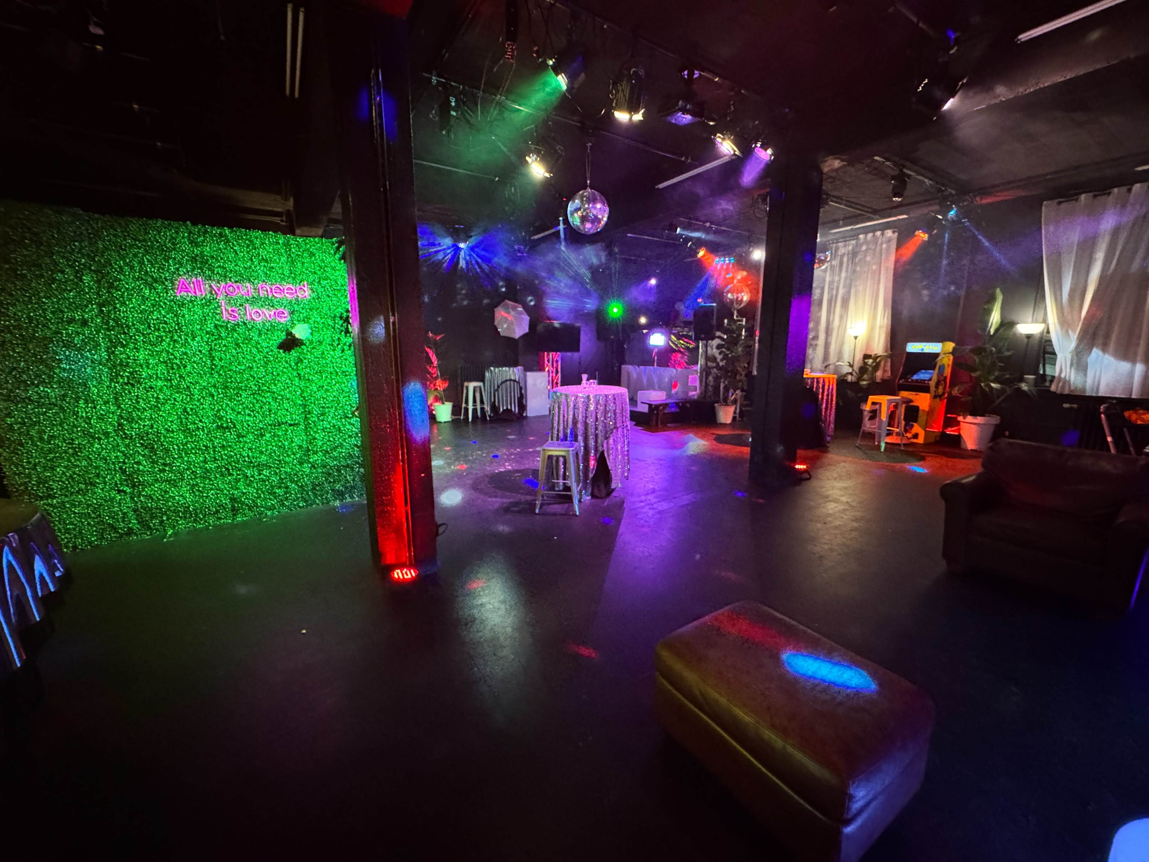 The image shows a dimly lit event space with colorful lights, a green foliage backdrop displaying the phrase "All You Need Is Love," and various seating arrangements.