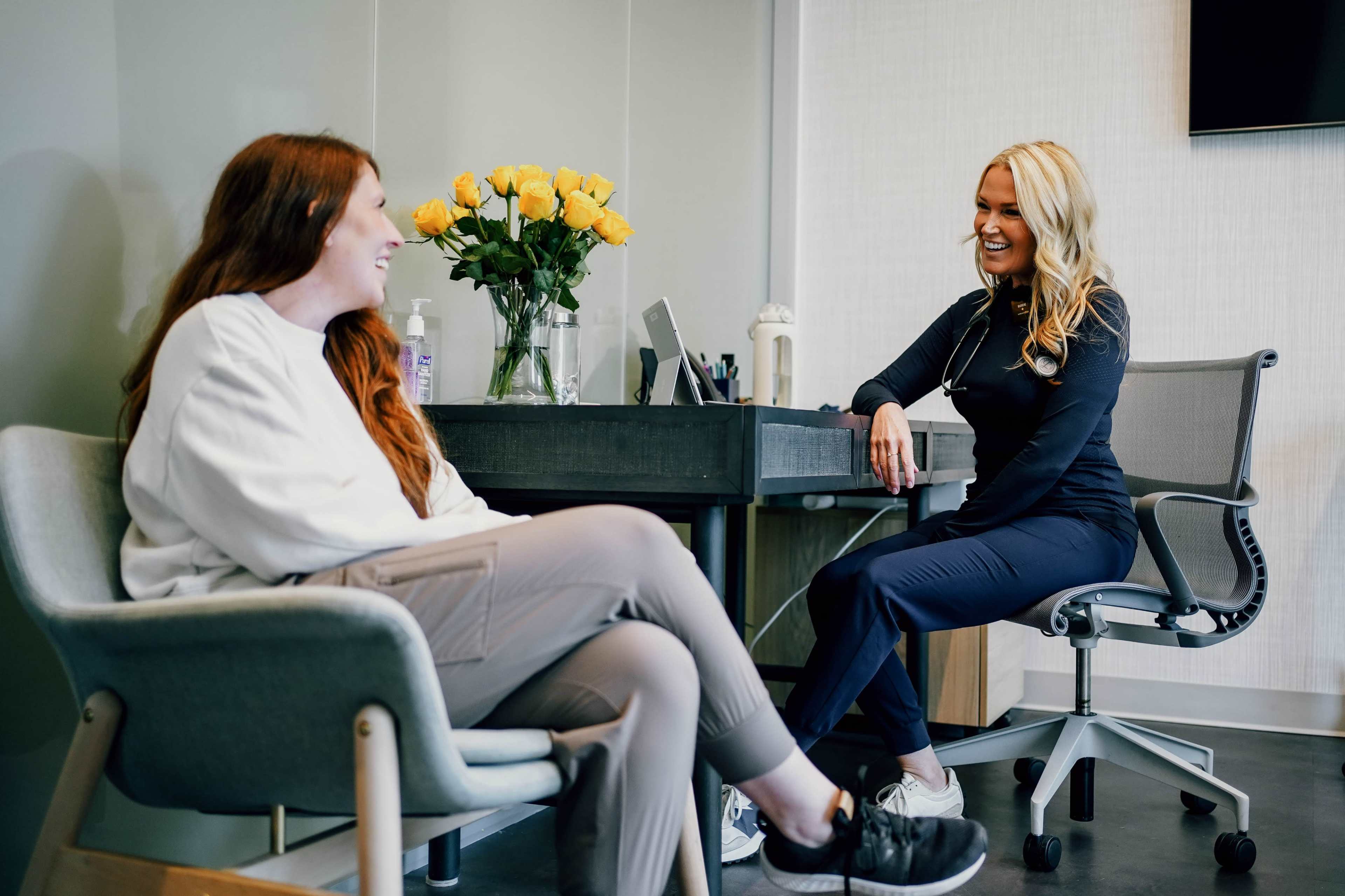 A healthcare professional sits across from a patient in a modern office, engaged in conversation, with a vase of yellow roses on the desk beside them.