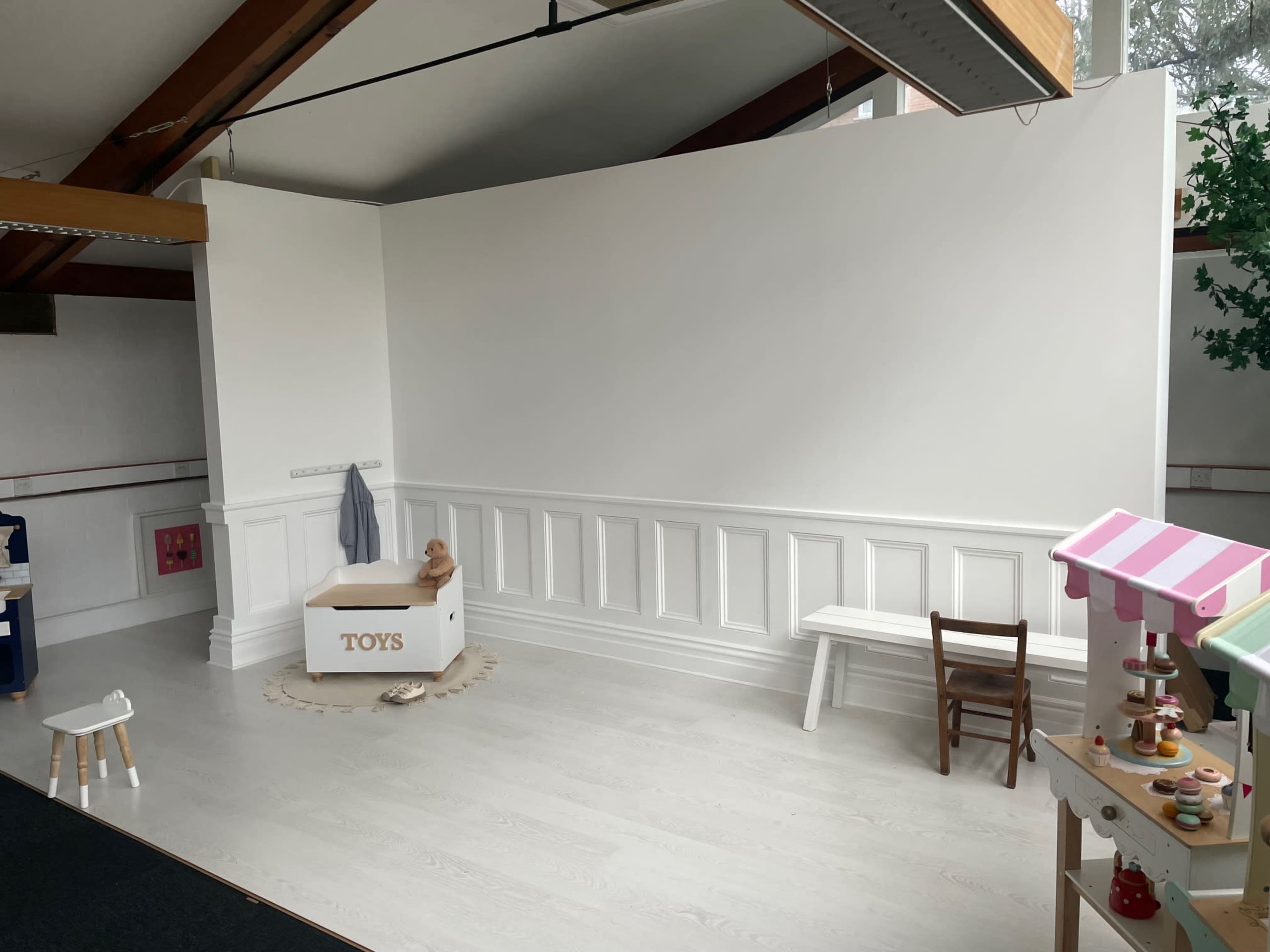 A minimalist play area featuring a white wall, a toy box labeled "TOYS," a small chair, and a few decorative elements on the floor.