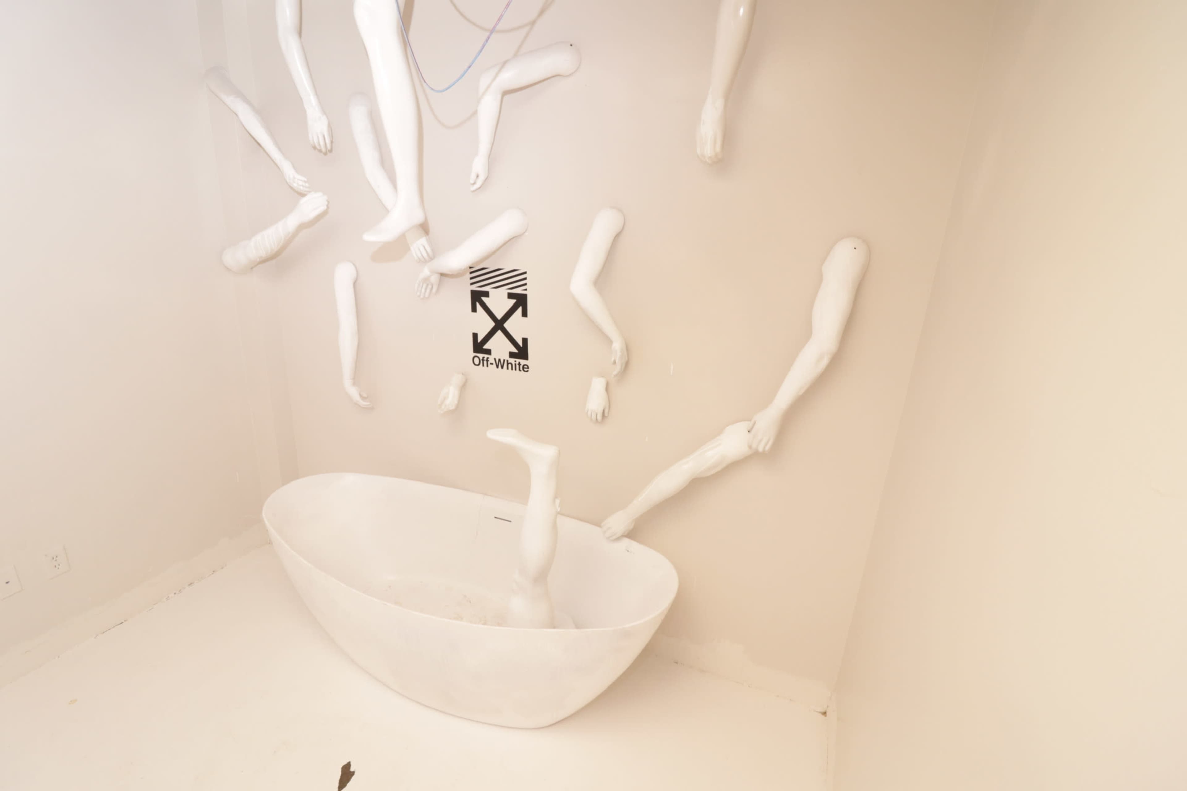 A white bathtub surrounded by multiple white, abstract arm sculptures mounted on a white wall.