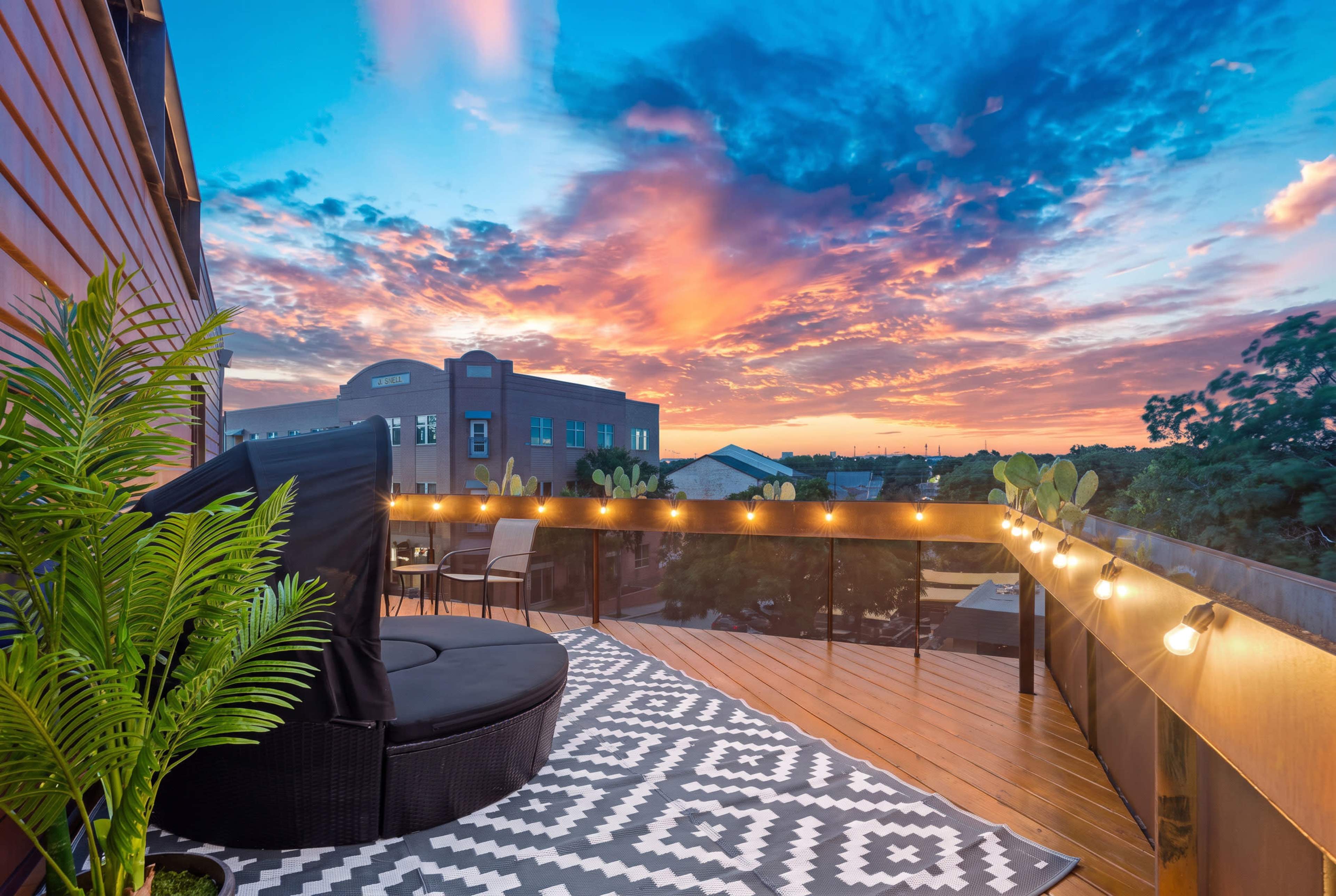 A balcony features a large black chair and potted plants, with a patterned rug and string lights illuminating the space, against a backdrop of a vibrant sunset sky.