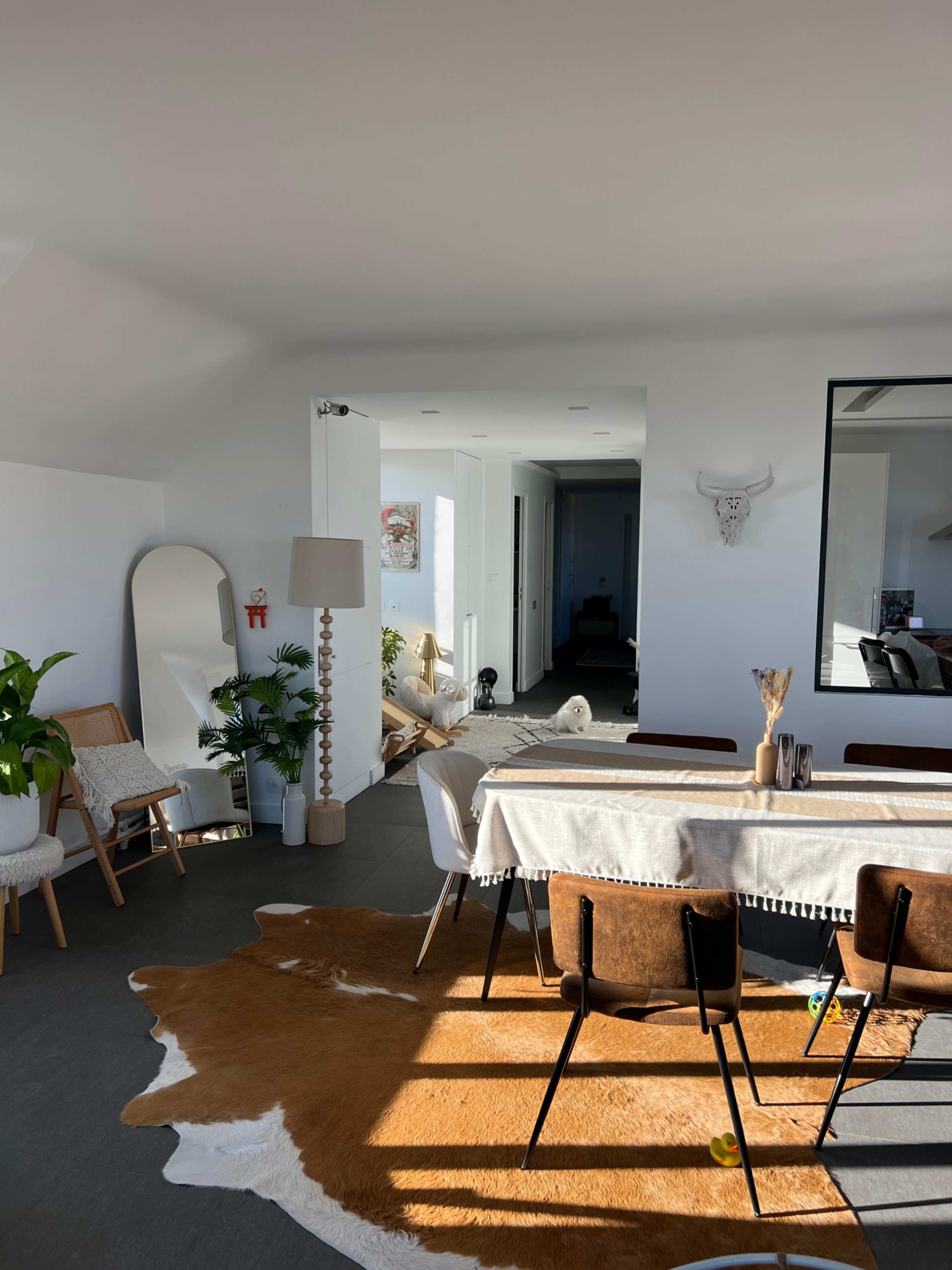 The image shows a brightly lit, modern interior featuring a dining area with a table set for meals, surrounded by chairs, a decorative cowhide rug, and plants, with an adjacent hallway and living space visible.