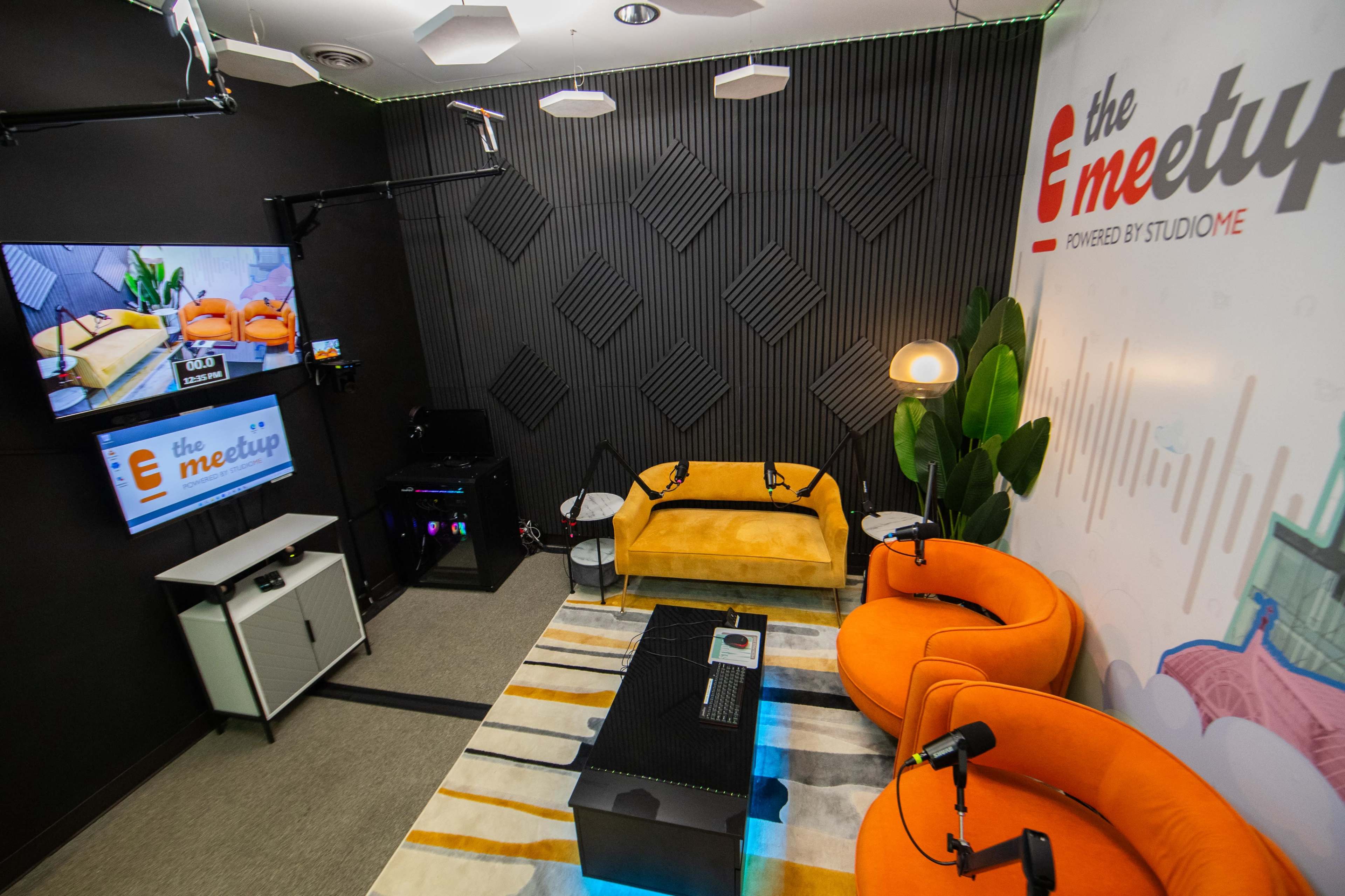 A modern recording studio with two orange chairs, a yellow couch, a striped rug, and a television mounted on the wall displaying the logo "the meetup."