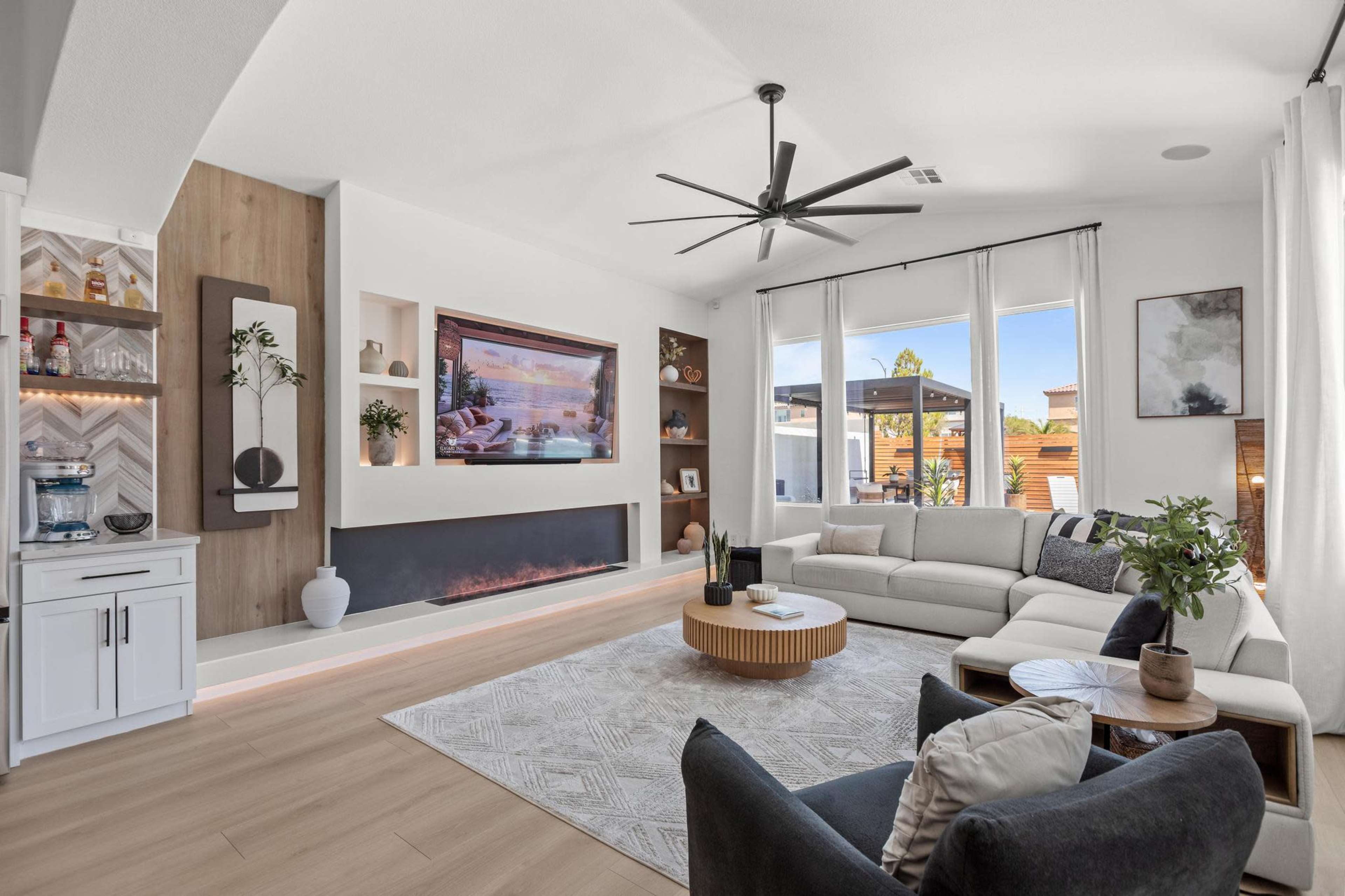 The image shows a modern living room with a large sectional sofa, a circular coffee table, a wall-mounted television, and large windows allowing natural light to enter.