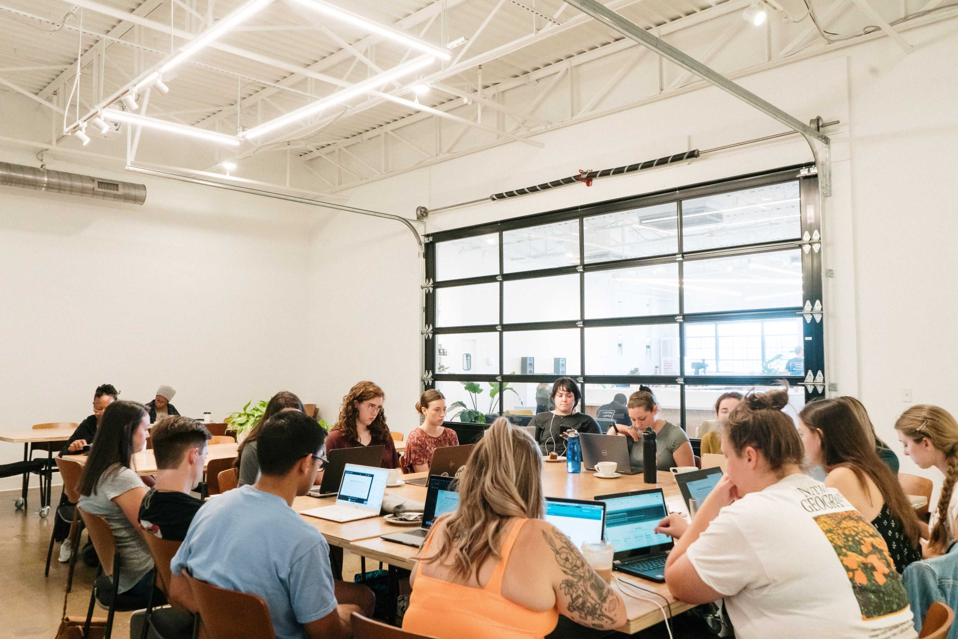 A group of people works at laptops around a large, communal table in a well-lit room with a large garage-style glass door.