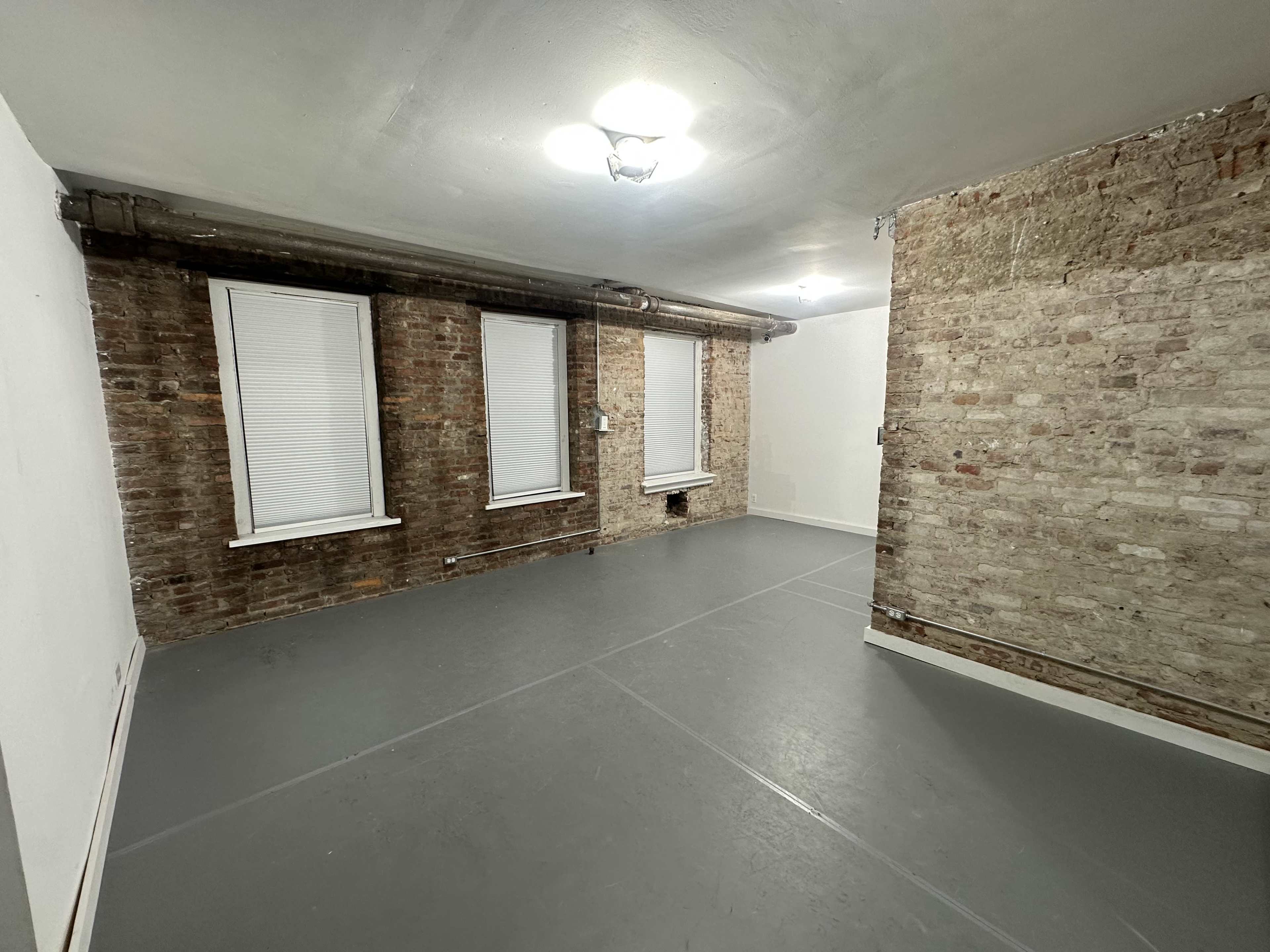 A sparsely furnished room with exposed brick walls, three windows with blinds, and a gray floor.