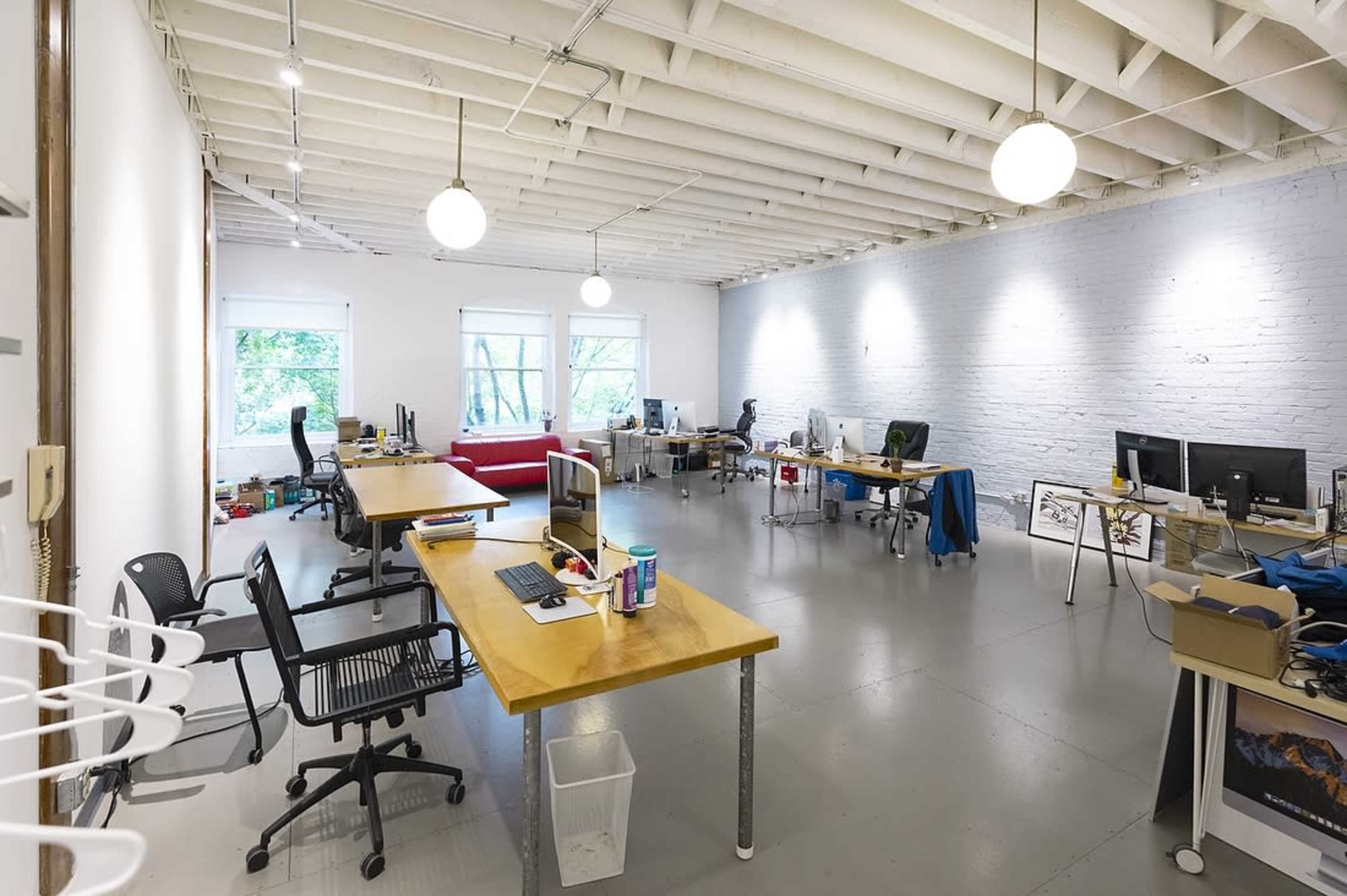 A spacious office with several desks, computers, and a red sofa, illuminated by overhead lights and large windows.