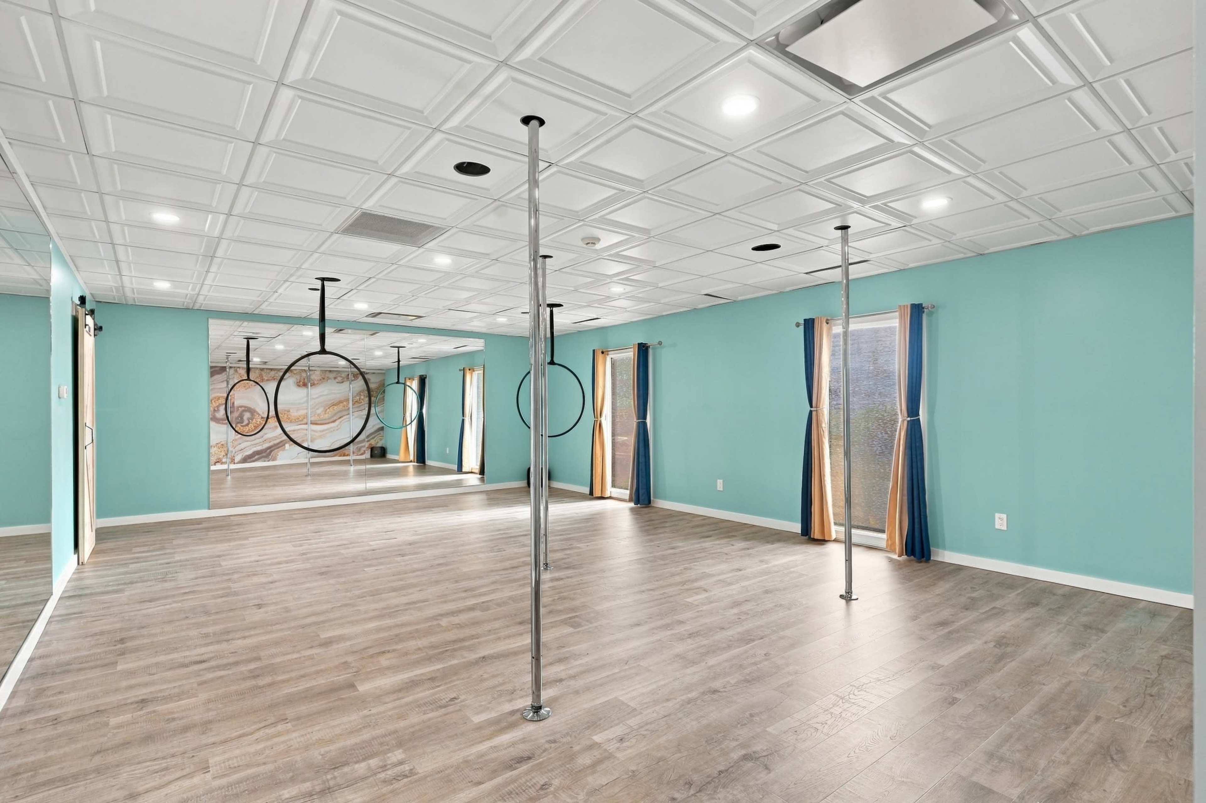 The image shows an empty room with turquoise walls, large mirrors, wooden flooring, and poles for aerial exercises.