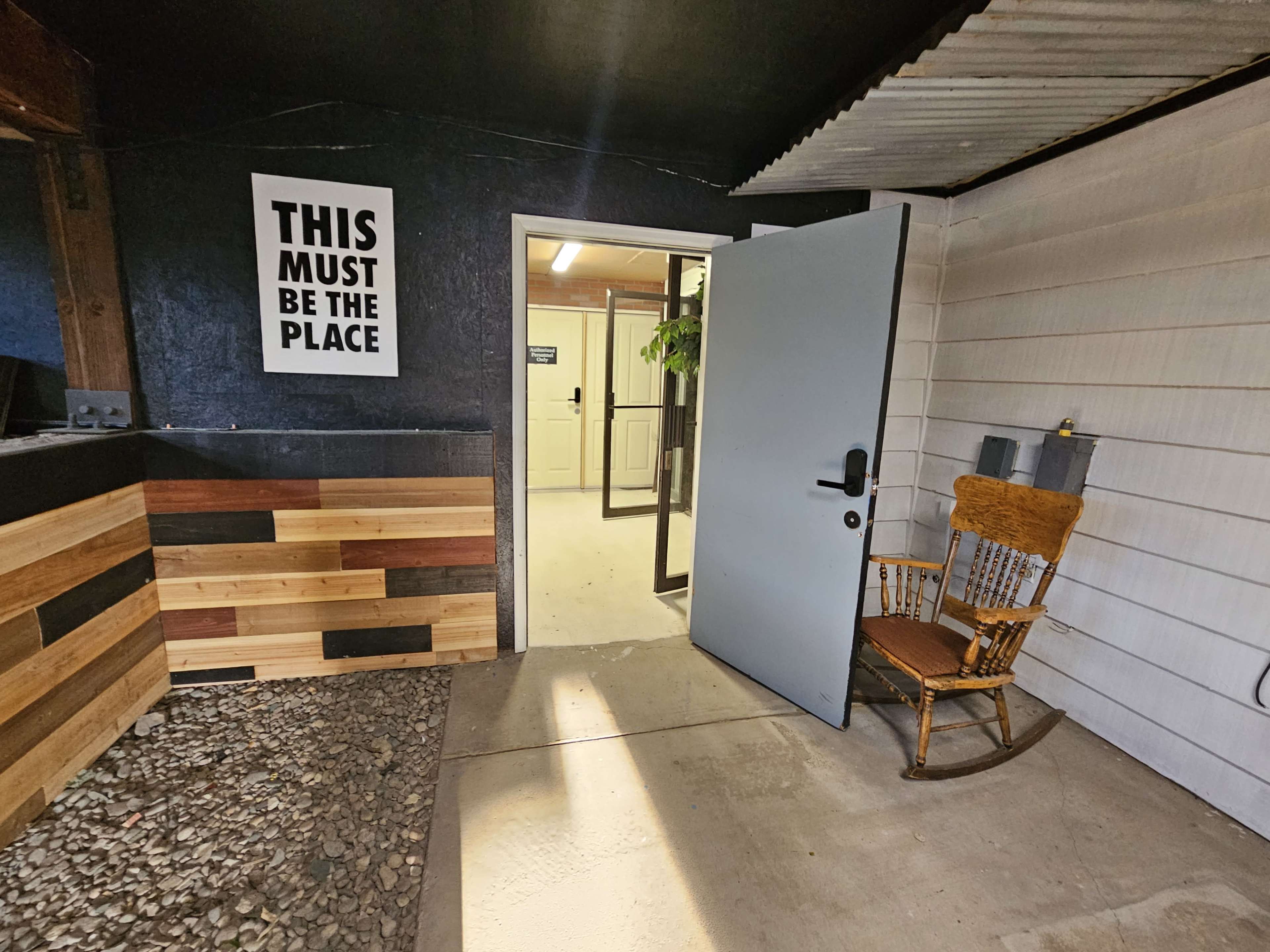 A doorway leading into a room with a sign that reads "THIS MUST BE THE PLACE," a rocking chair beside the entrance, and a wooden wall with various colored panels.